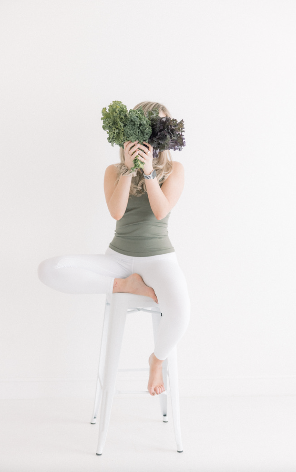 Woman sitting on a stool with one leg crossed, holding a bunch of leafy greens in front of her face against a white background