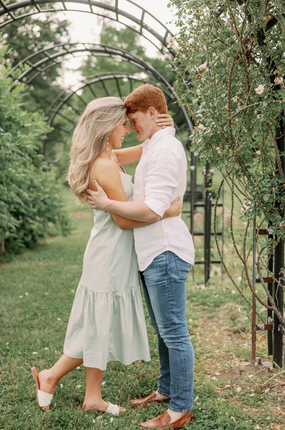 A couple embracing in a garden with green foliage and archways, faces close together, smiling.