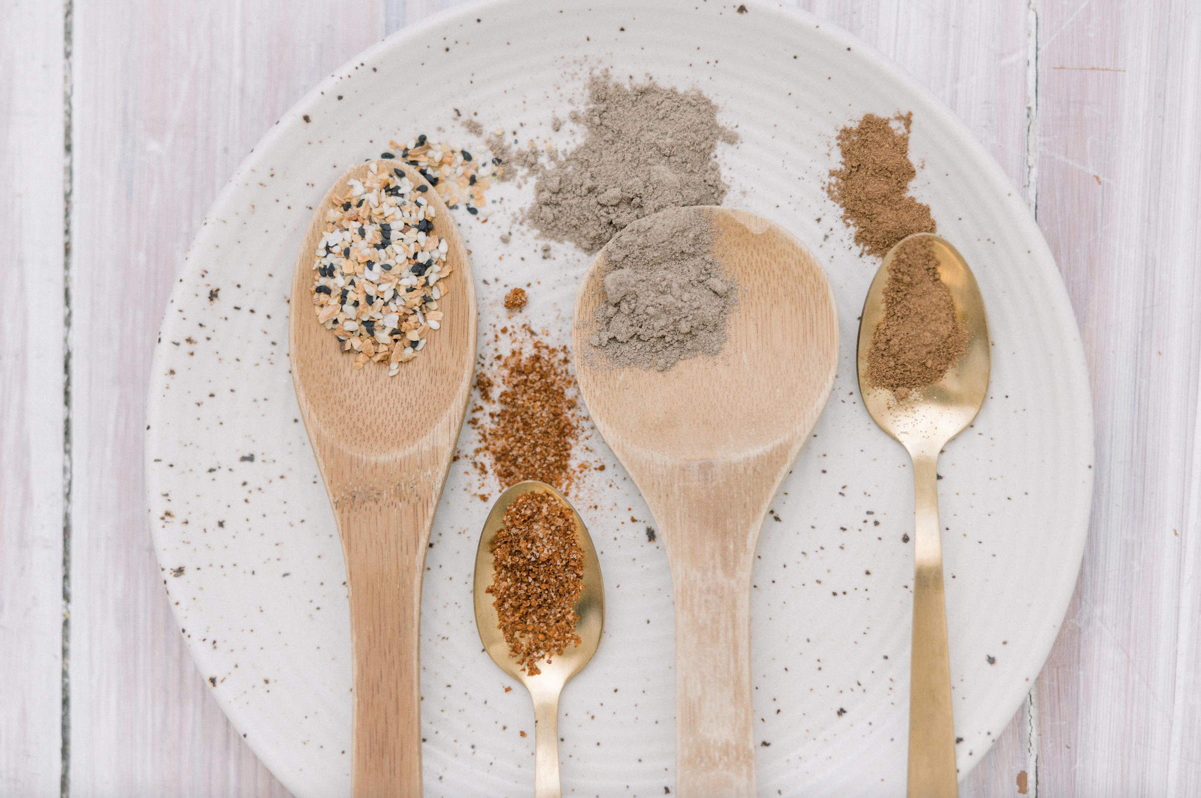 Multiple types of ground spices in small spoons on a white speckled ceramic plate, arranged in a row, on a light wooden surface.