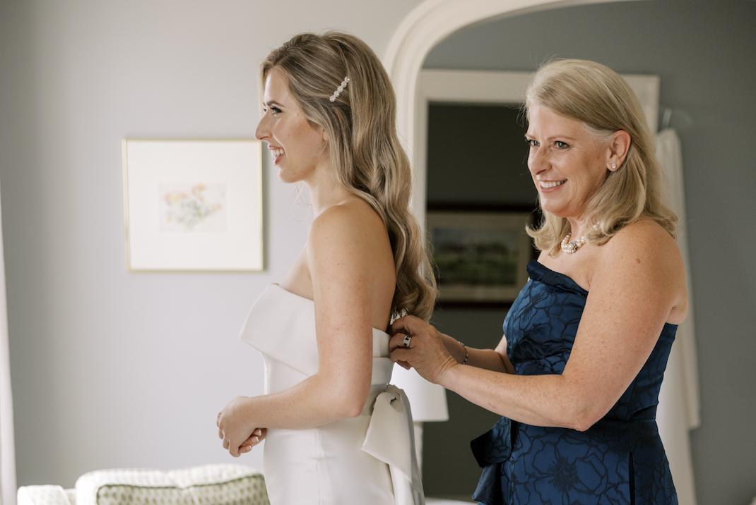A smiling young woman in a white strapless wedding dress with a bow at the back is being assisted by an older woman in a blue dress as she gets ready for her wedding.