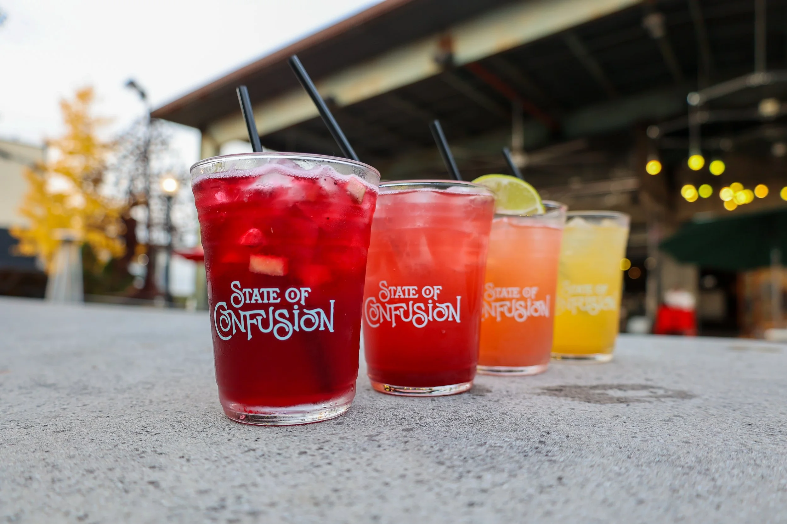 Four colorful drinks in glasses with black straws, each with a different garnish, on a gray table outdoors under a structure, with blurred trees and lights in the background.