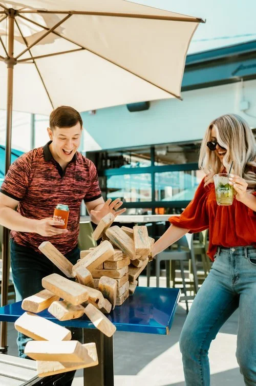 A young man and woman playing a game of Jenga outdoors at a cafe or restaurant patio, with a large umbrella and glass windows in the background, both smiling and having fun.