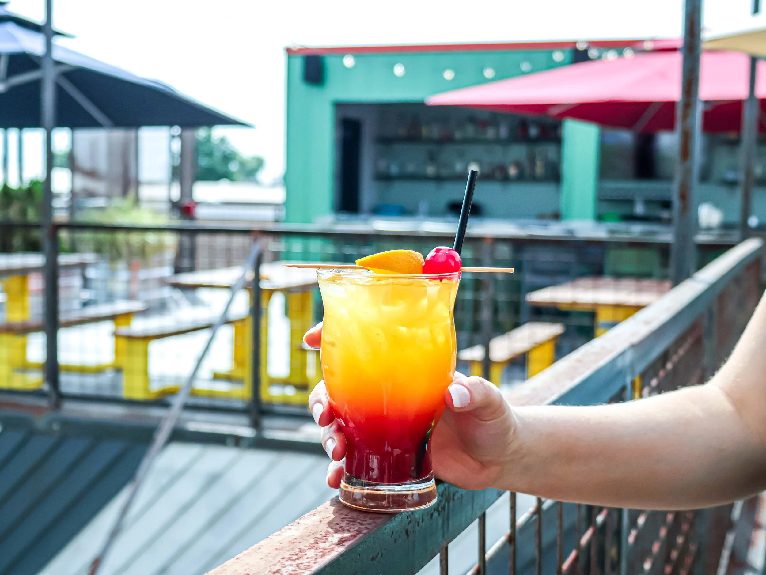 A hand holding a colorful layered tropical cocktail with fruit garnish on a balcony with outdoor seating and a green building in the background.