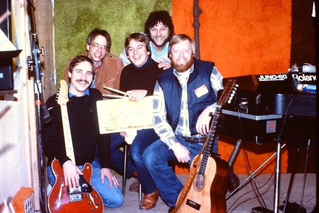 Six people in a musical studio, some holding guitars and a keyboard, smiling for the photo.