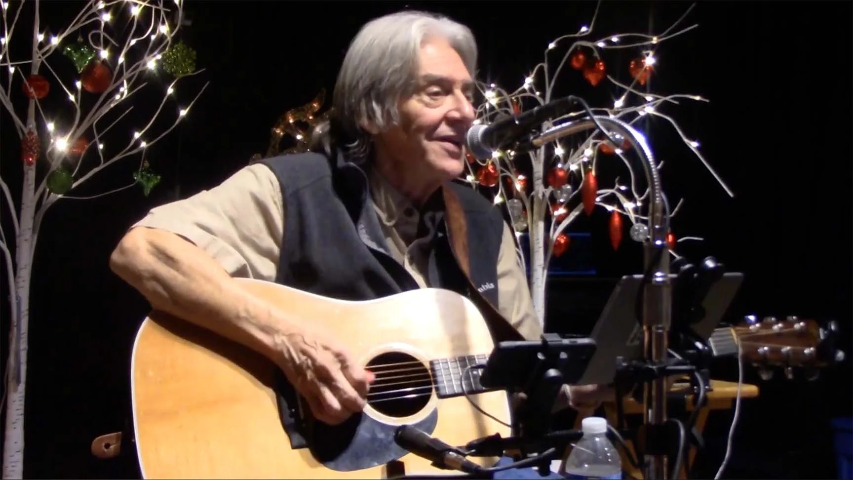 An older man with gray hair playing an acoustic guitar and singing into a microphone during a festive event, with decorative holiday lights and ornaments in the background.