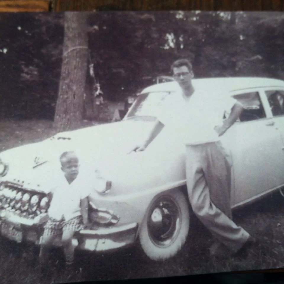 A black and white photo of two people with a vintage car outdoors, likely in a wooded area.
