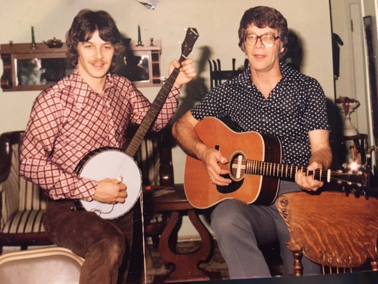 Two men sitting indoors, playing guitars; the man on the left is holding a banjo, and the man on the right is playing an acoustic guitar. They are smiling and looking at the camera.