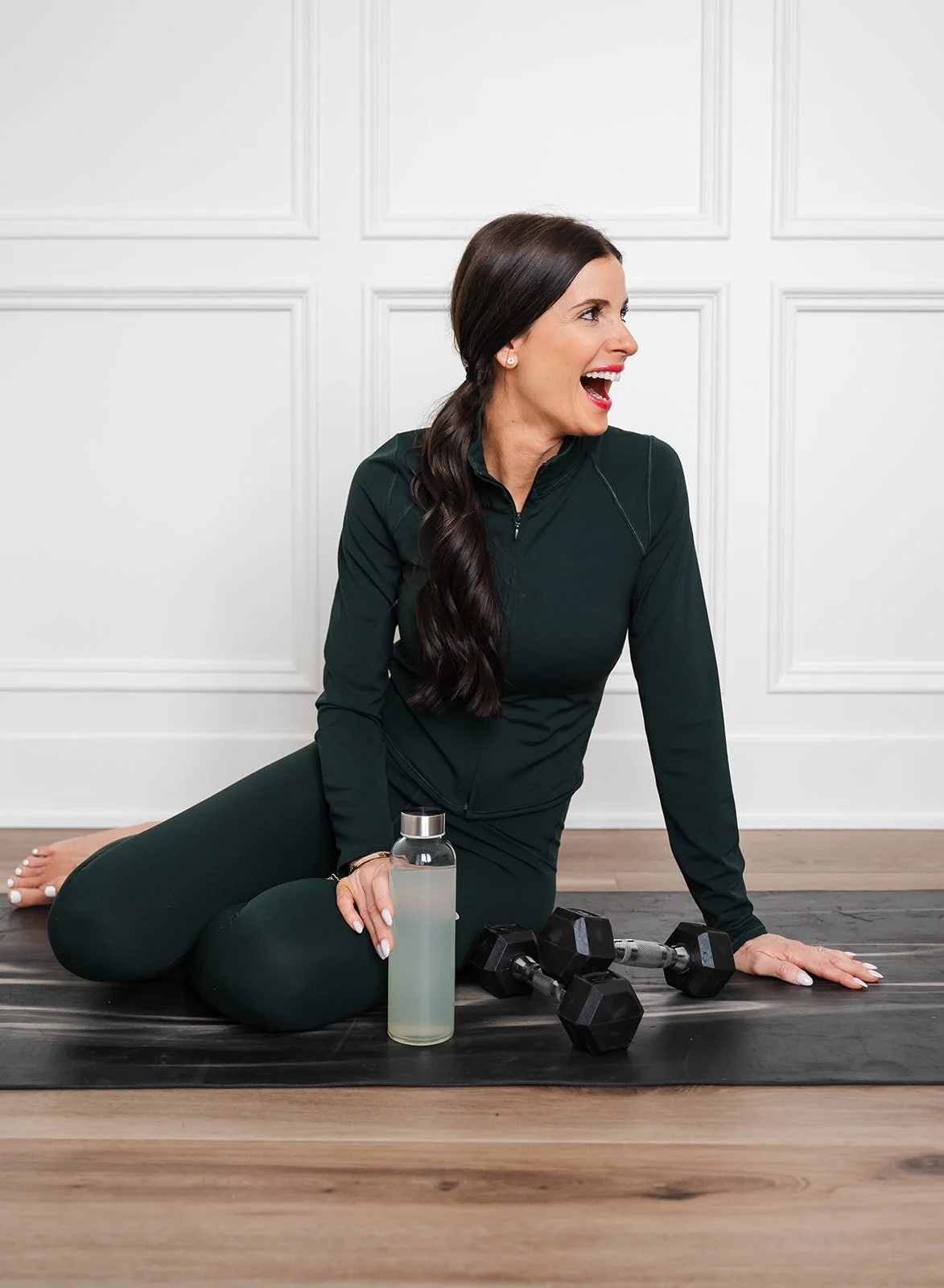A woman with long dark hair and a dark green athletic outfit is sitting on a black yoga mat on a wooden floor, smiling and looking to her right. She has two dumbbells, a water bottle, and a skincare or water spray bottle in front of her.