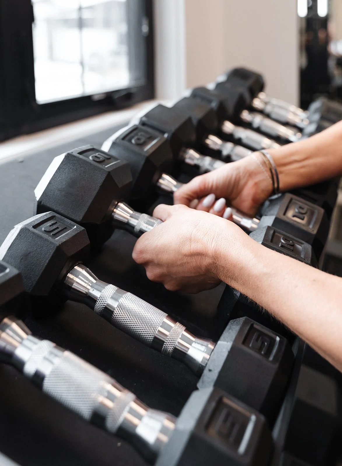 Person adjusting dumbbells on a rack at the gym with a window in the background.