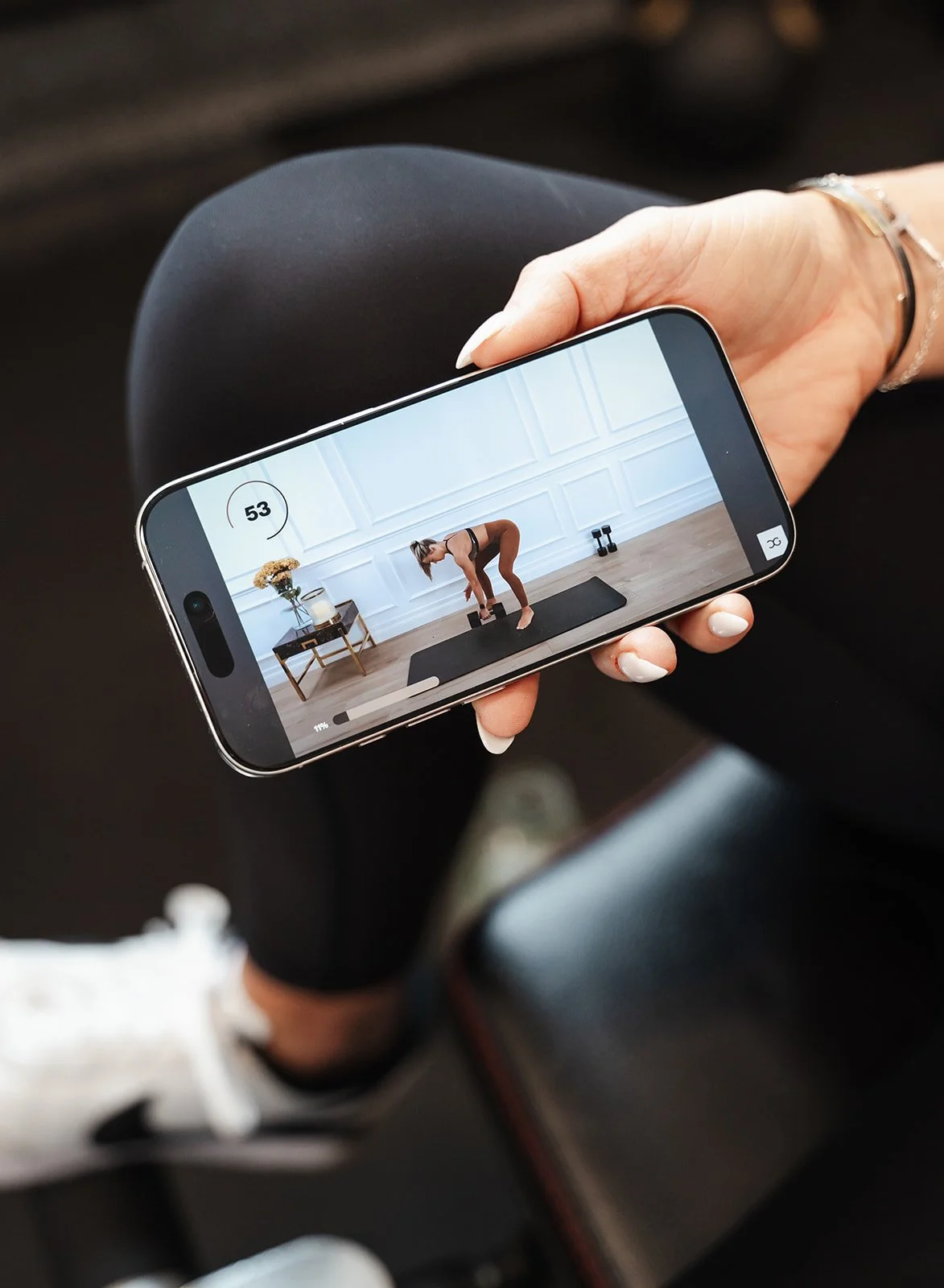A person is taking a photo or video of a woman exercising with dumbbells on a yoga mat in a white paneled room, using a smartphone.