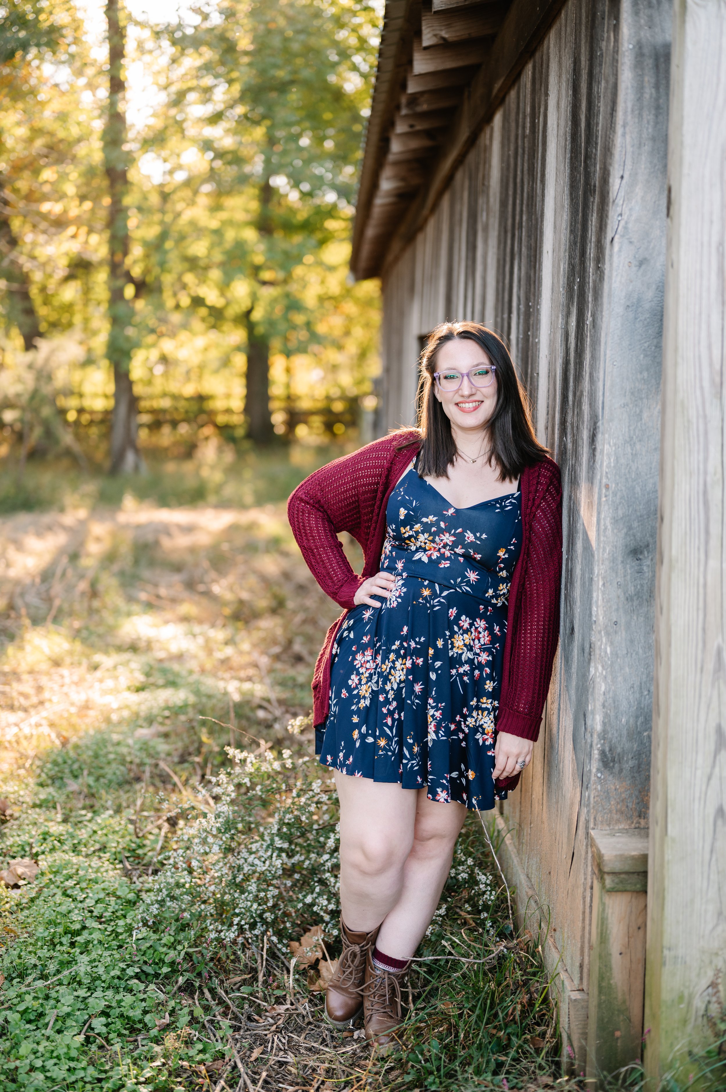 A woman with dark hair, wearing glasses, a navy floral dress, a red cardigan, and brown boots, leaning against a wooden wall in a grassy area with trees in the background during sunset.