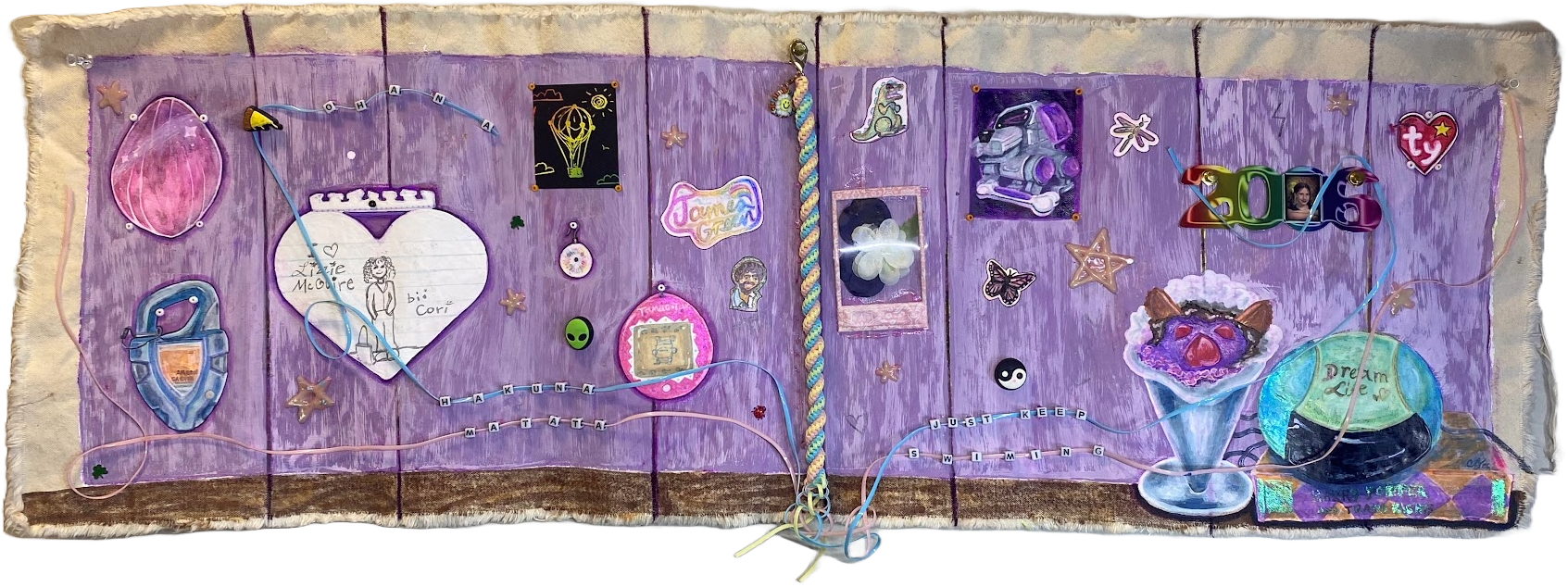 A colorful, decorated fabric wall hanging with various inspirational and whimsical items, including drawings, a rainbow-colored rope, small figures, pictures, and themed embellishments, with a purple background and a brown bottom edge.