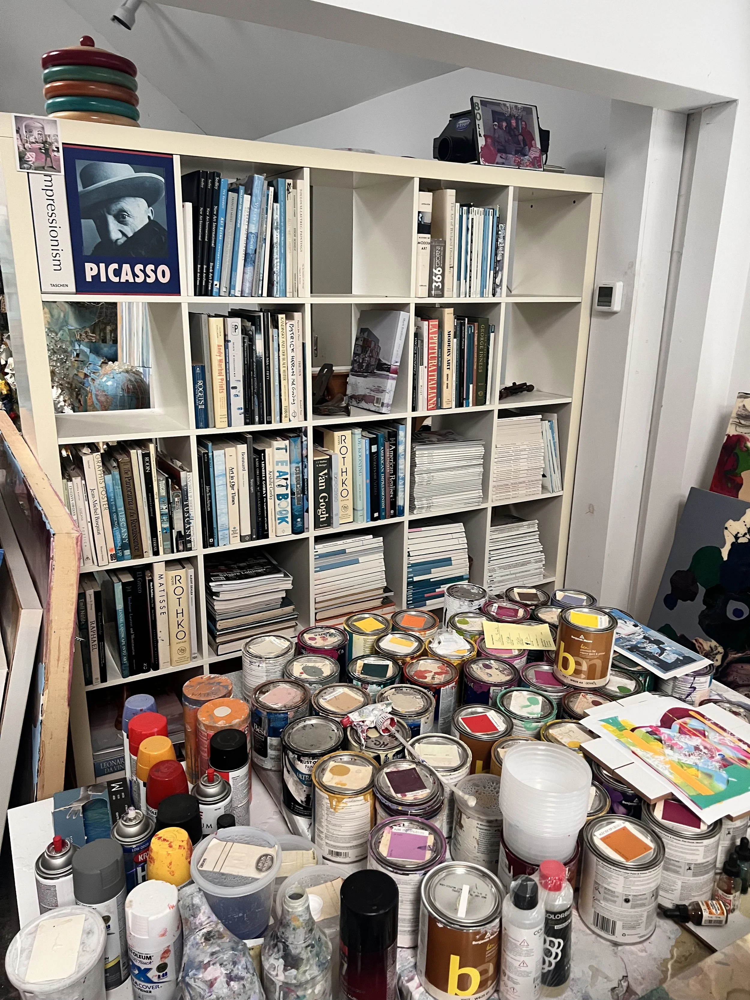 A cluttered art studio with shelves filled with books, art supplies, and a table covered with paint cans, spray cans, and art materials.