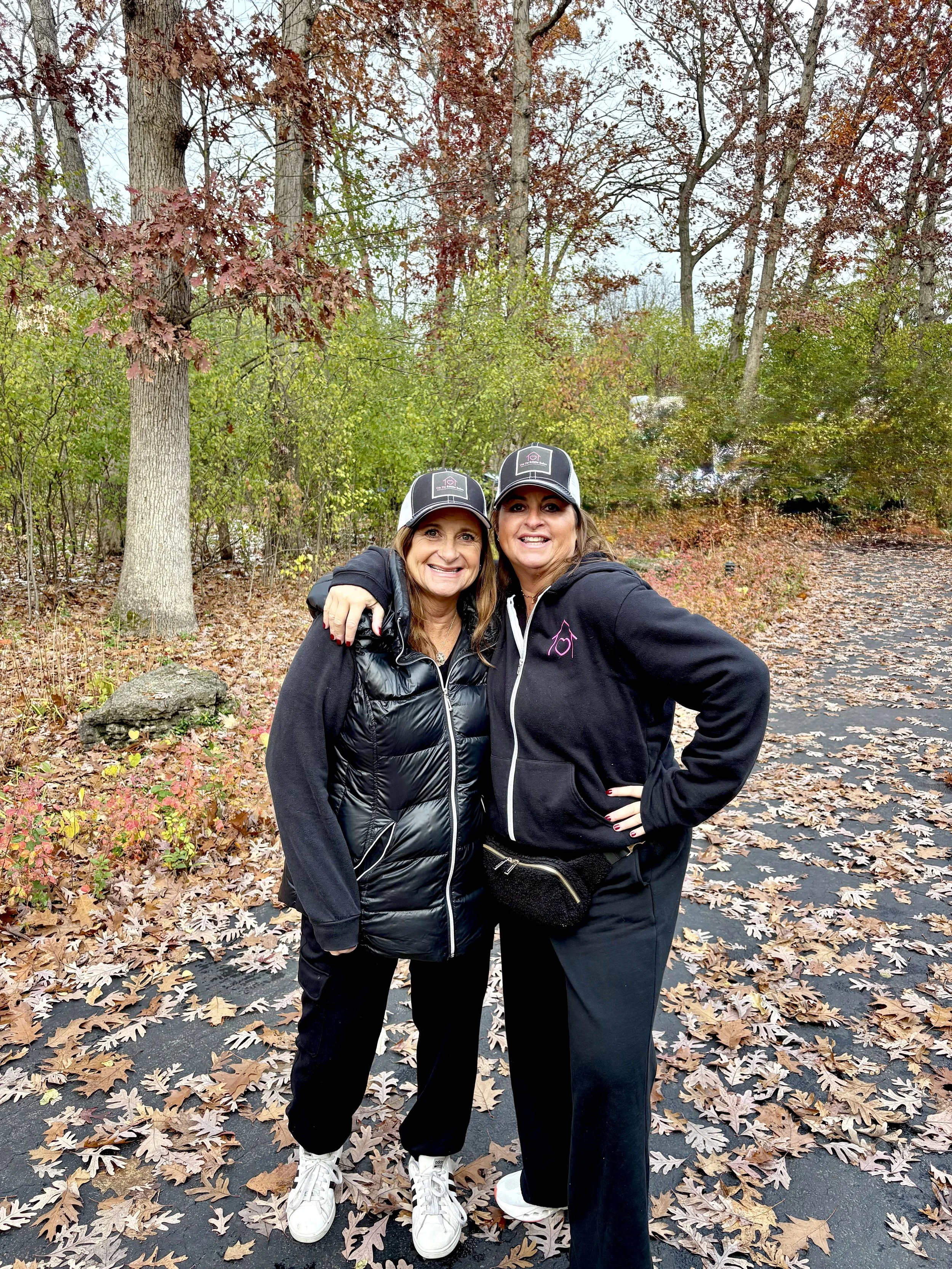 Two women in black jackets and black pants standing outdoors on a fall day, smiling and hugging each other, with a background of trees with autumn leaves and a leaf-covered ground.