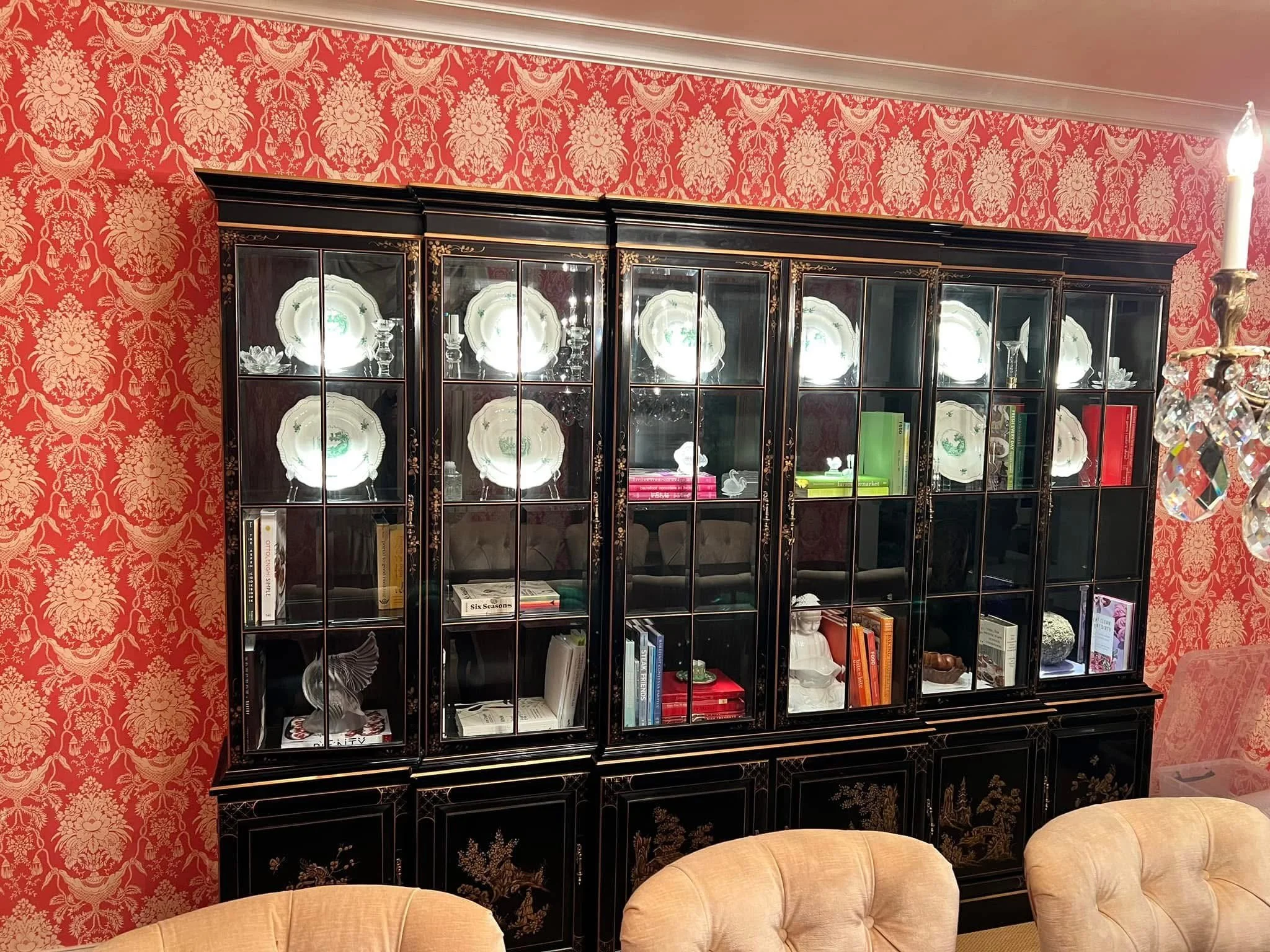 A black china cabinet with gold details on a red damask wallpaper background. The cabinet glass doors display decorative plates and various books, with a tan upholstered chair in the foreground and a chandelier with crystals on the right side.
