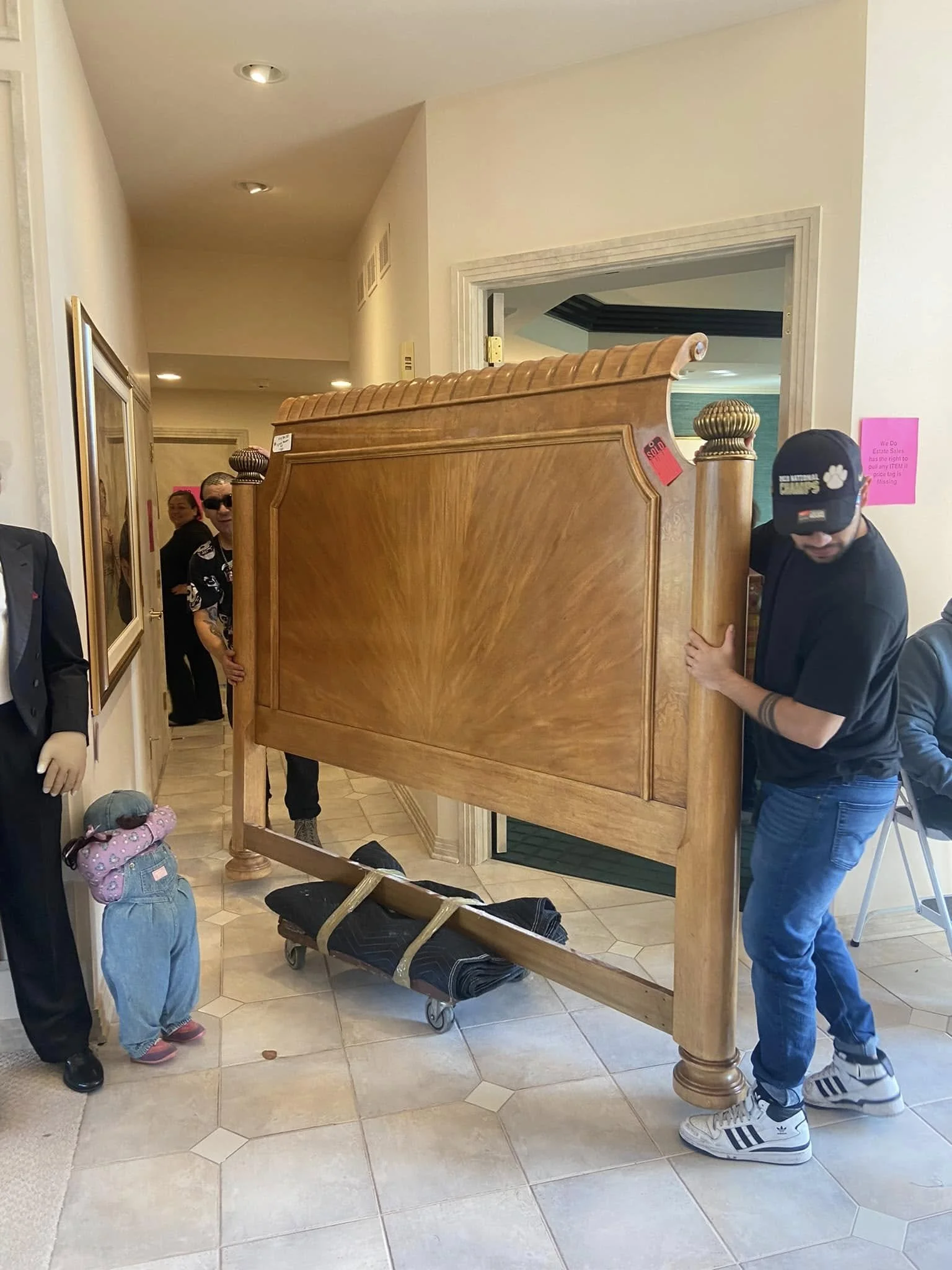 Two men are moving a large wooden piece of furniture, possibly a headboard, through a doorway. One man is wearing a black t-shirt and sneakers, while the other, wearing sunglasses, is partially hiding behind the furniture. There are two people in the