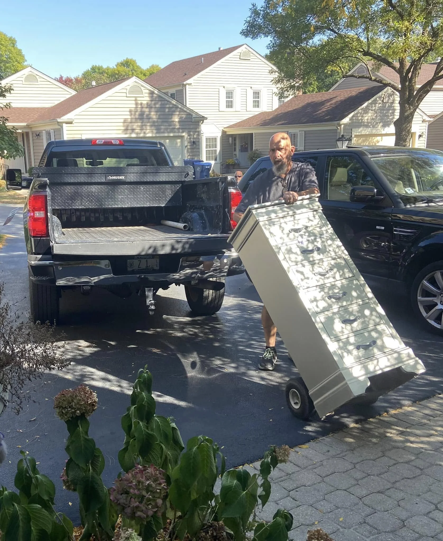 A man with a beard in a gray t-shirt and shorts loading a cream-colored dresser onto a black SUV in a suburban driveway. The driveway is shaded by trees, with houses and cars in the background.