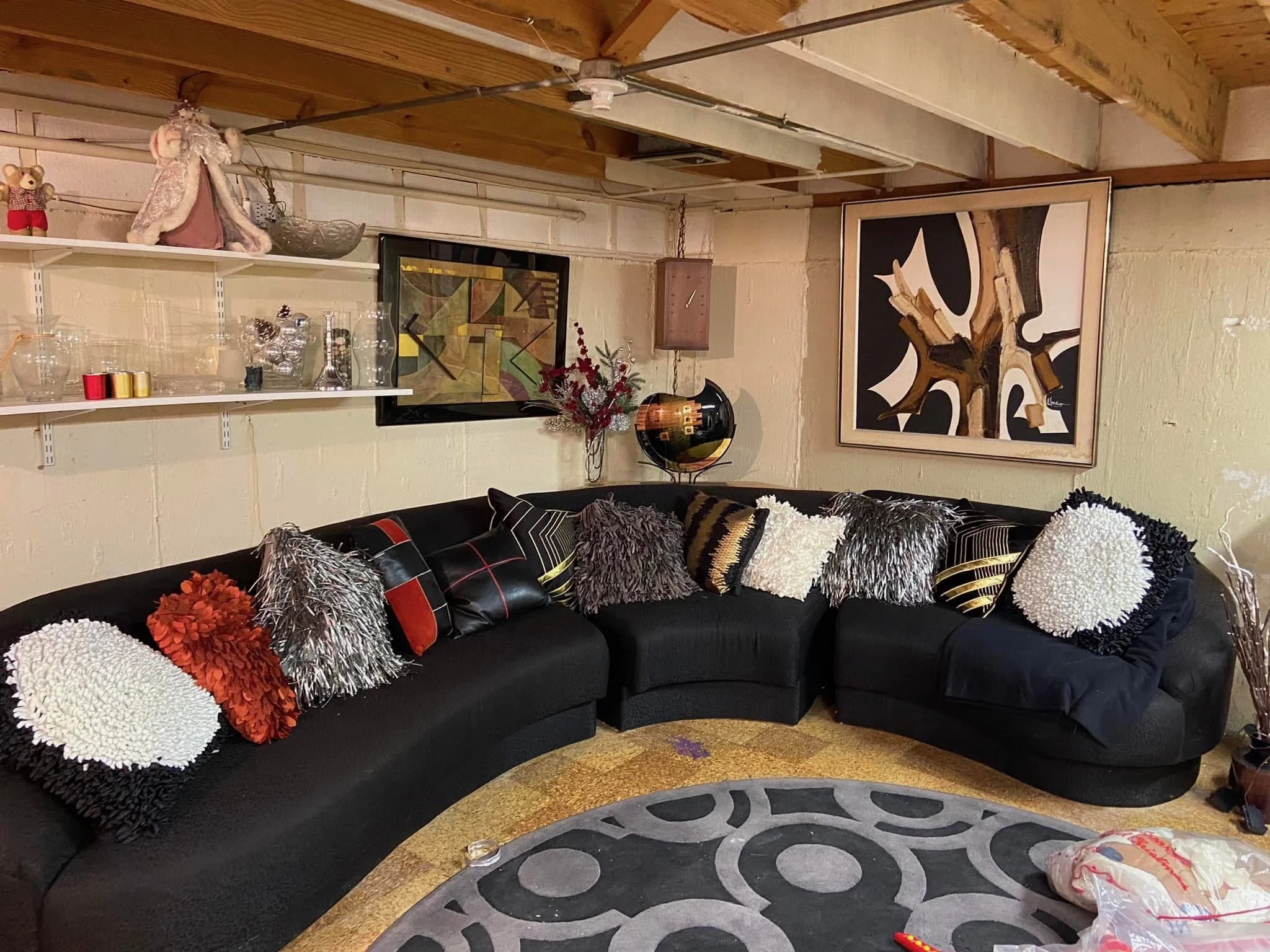 A sectional black sofa with colorful textured pillows, a patterned rug, wall art, decorative items on shelves, a vase with flowers, and a round table with a globe in a room with wooden ceiling beams and cream-colored brick walls.