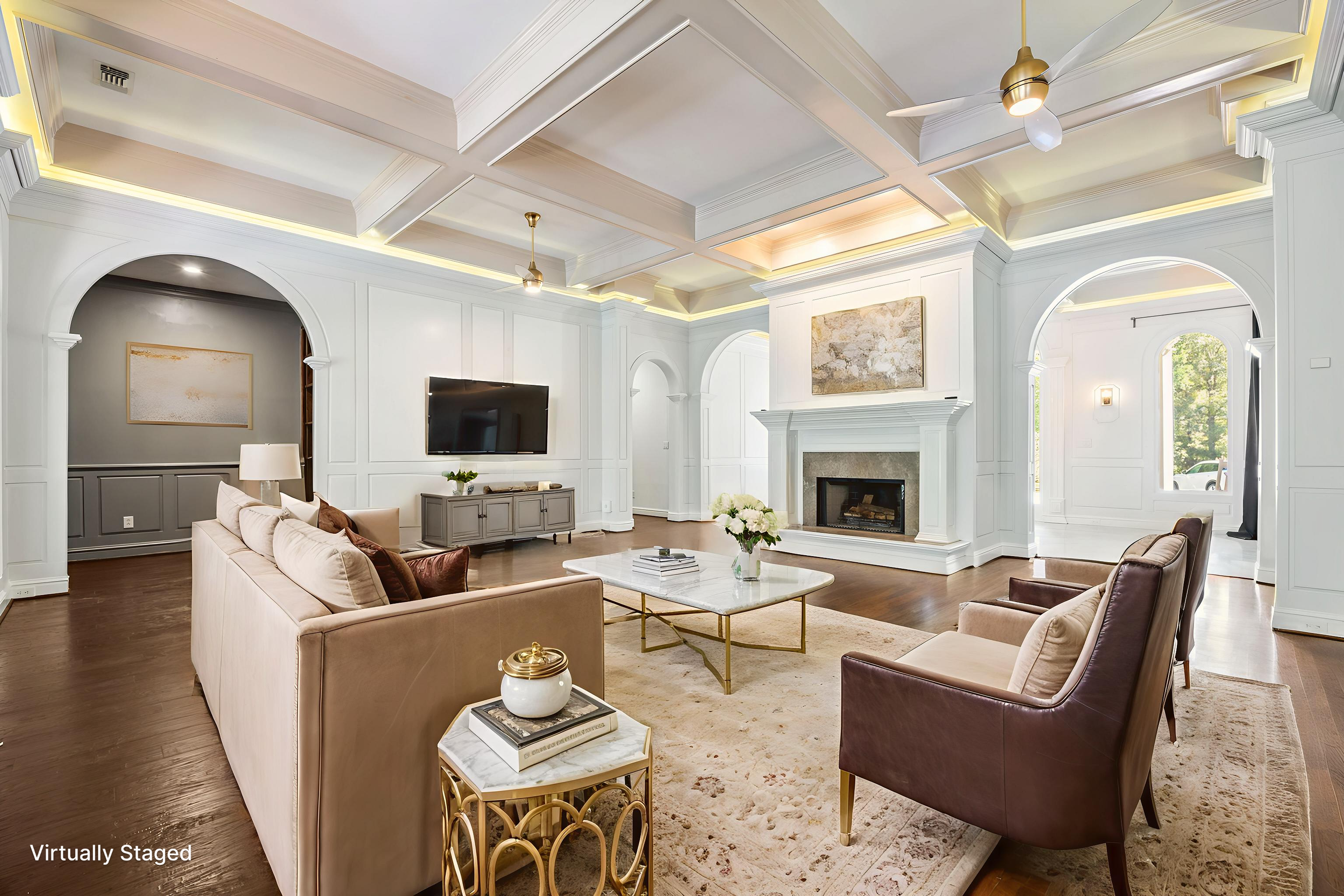 Elegant living room with white paneled walls, coffered ceiling with recessed lighting, a fireplace with white mantle and art above, a TV mounted on the wall, beige sofa, brown armchair, marble coffee table with flowers, and hardwood floors.