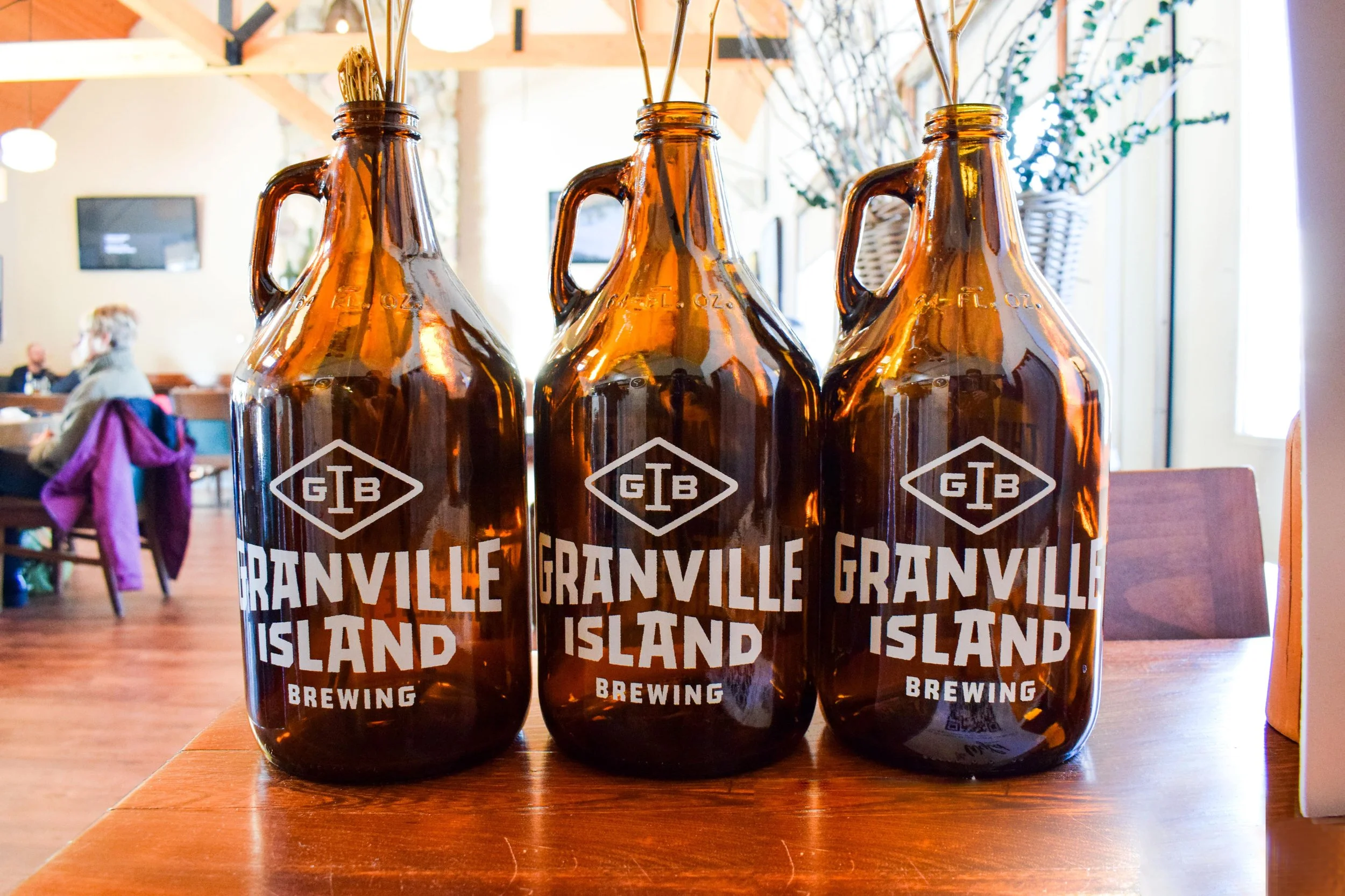 Three amber glass bottles with white text reading 'Granville Island Brewing' placed on a wooden table in a brightly lit indoor space, with blurred background of people and decor.