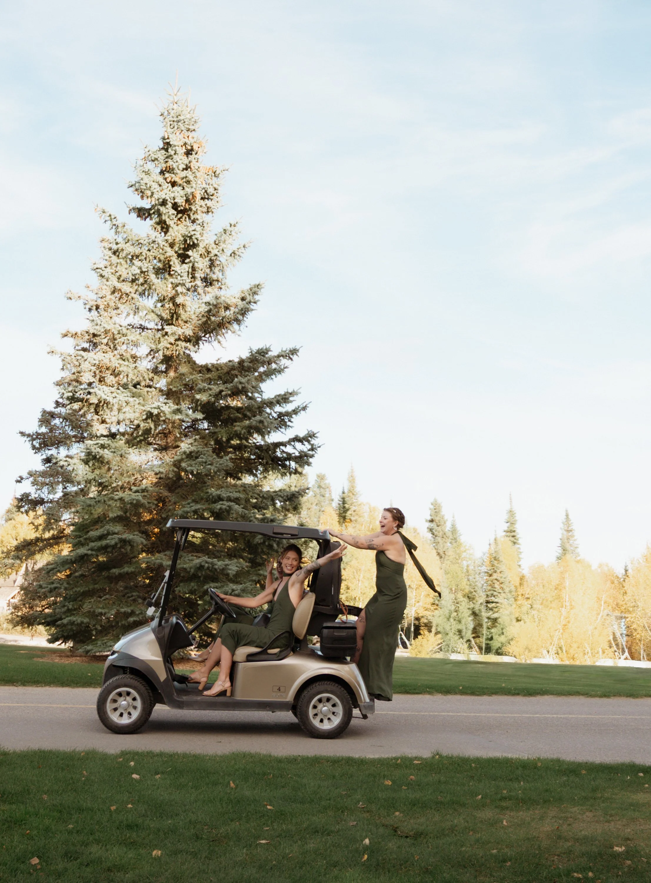 Two women in a green jumpsuit are riding in a golf cart on a paved path in a park, and a third woman in a green dress is standing beside the cart, holding one of the women, all smiling and enjoying themselves. There are trees and a lake in the background.
