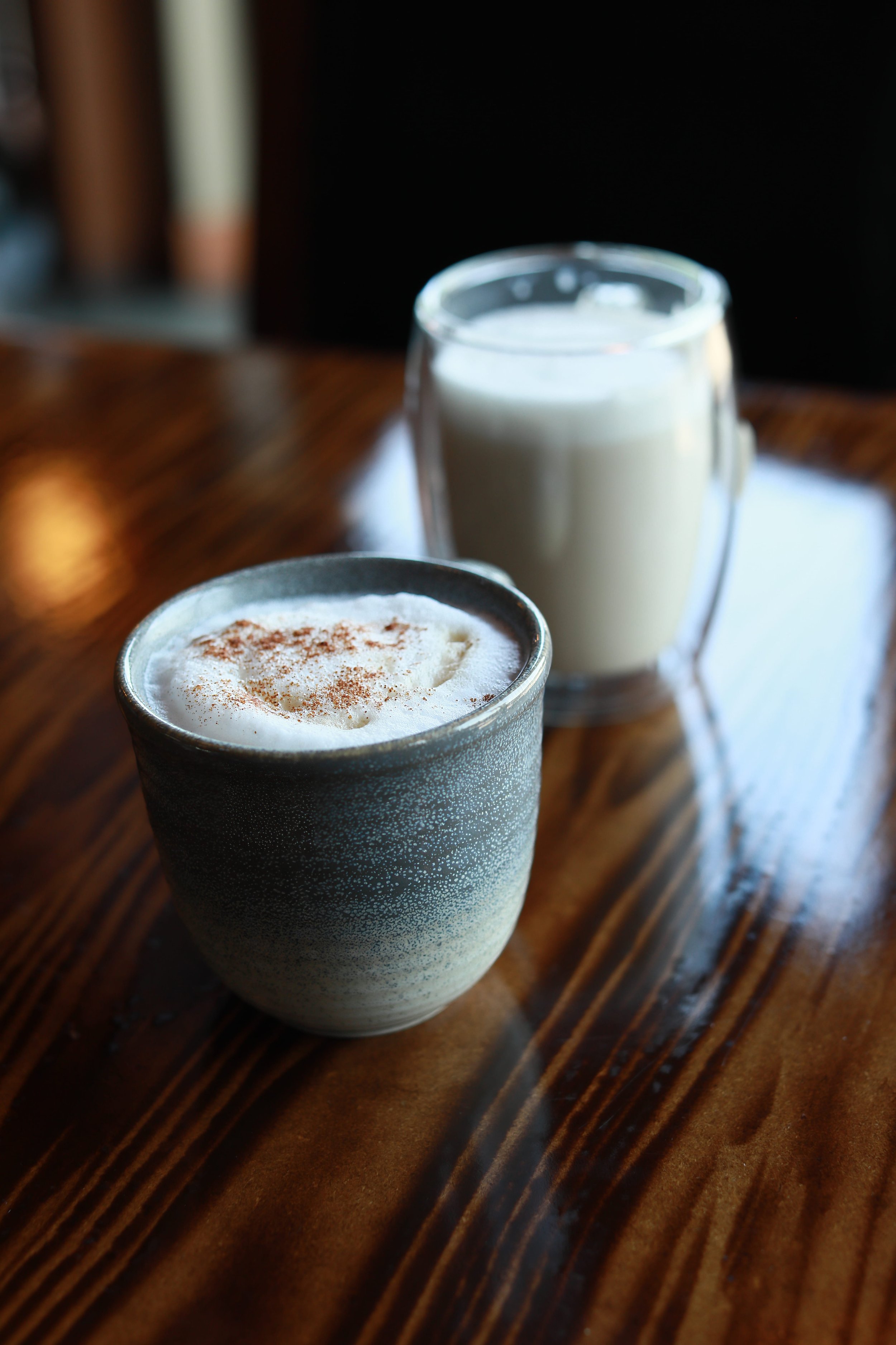 Close-up of two beverages on a wooden table, one is a hot drink topped with whipped cream and cocoa powder in a gray mug, the other appears to be a cold drink in a glass mug.