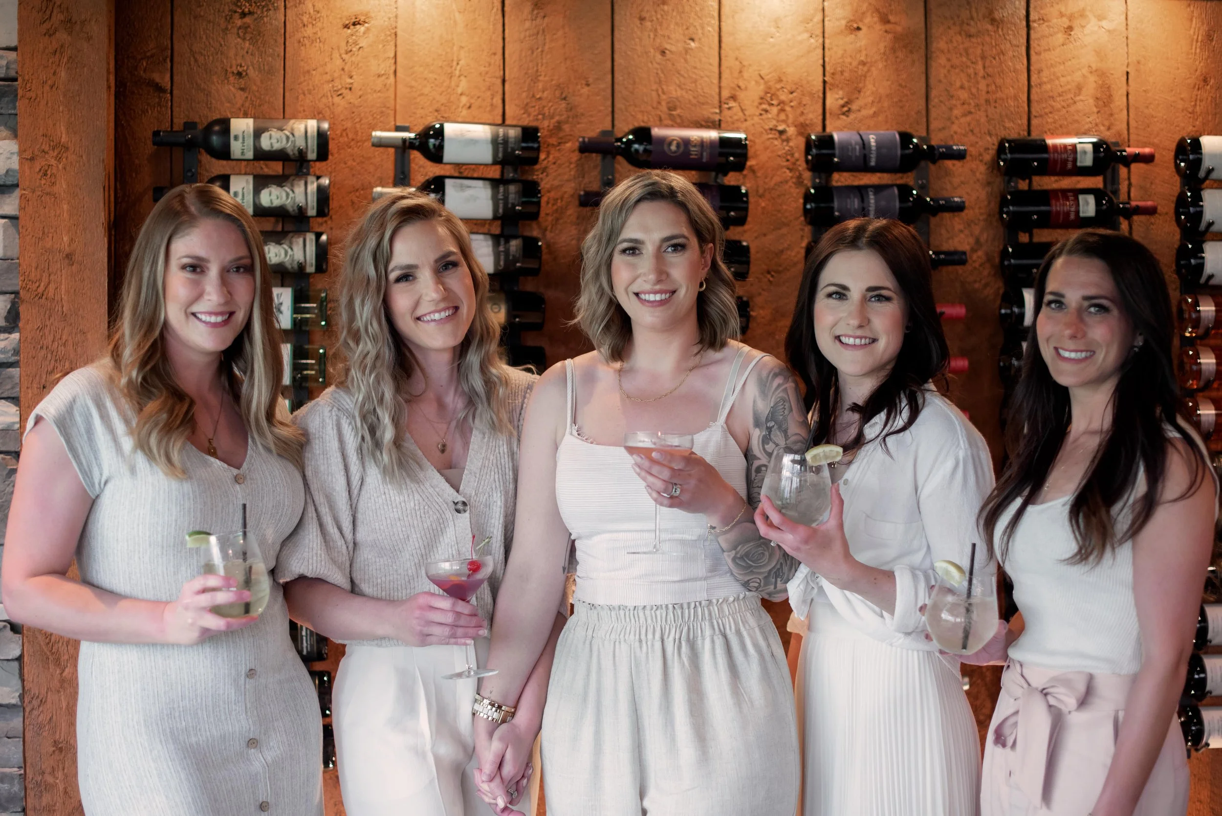 Six women in white dresses standing together at a bar or restaurant, holding drinks, with wine bottles on the wall behind them.