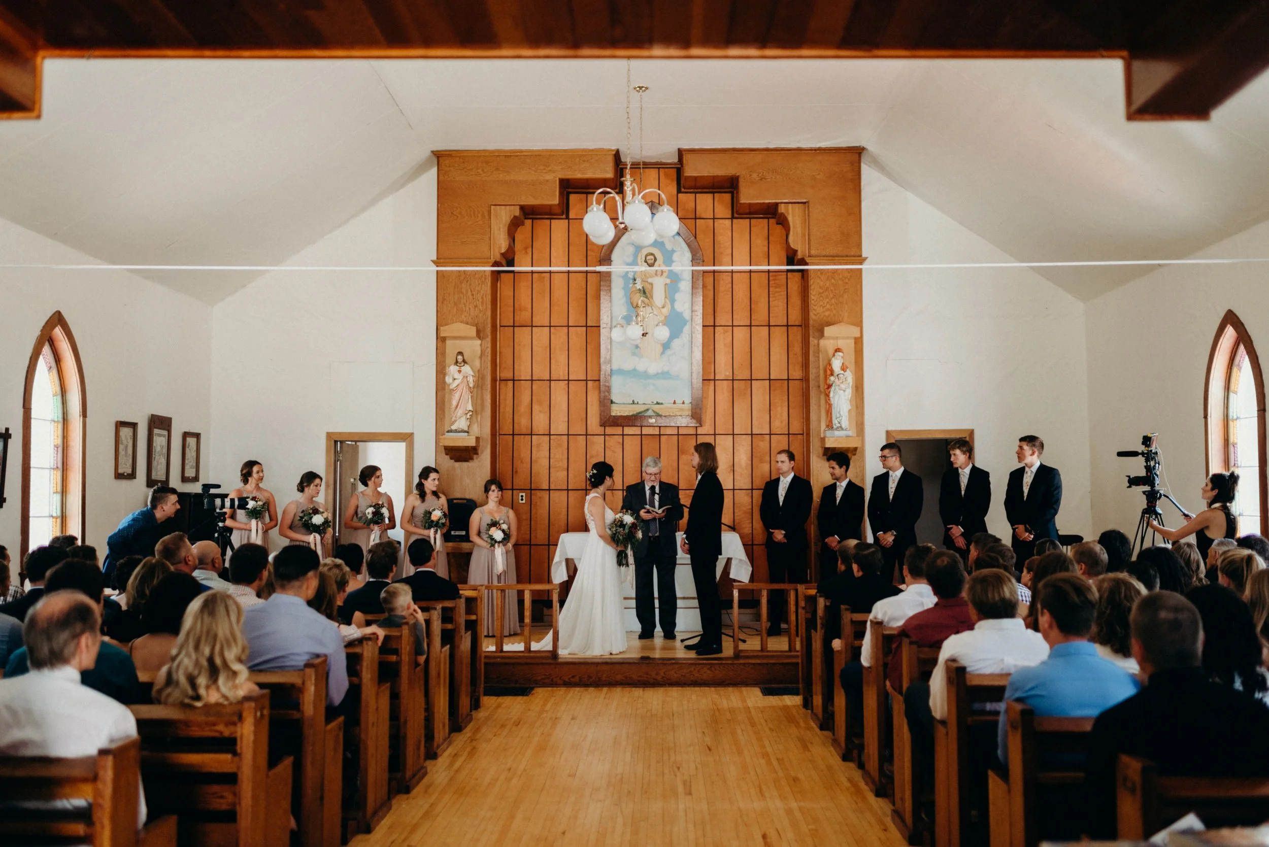 A wedding ceremony inside a church, with the bride and groom standing at the altar facing each other, surrounded by bridesmaids, groomsmen, and a priest. The church has wooden paneling and stained glass windows, with many guests seated in pews watching the ceremony.