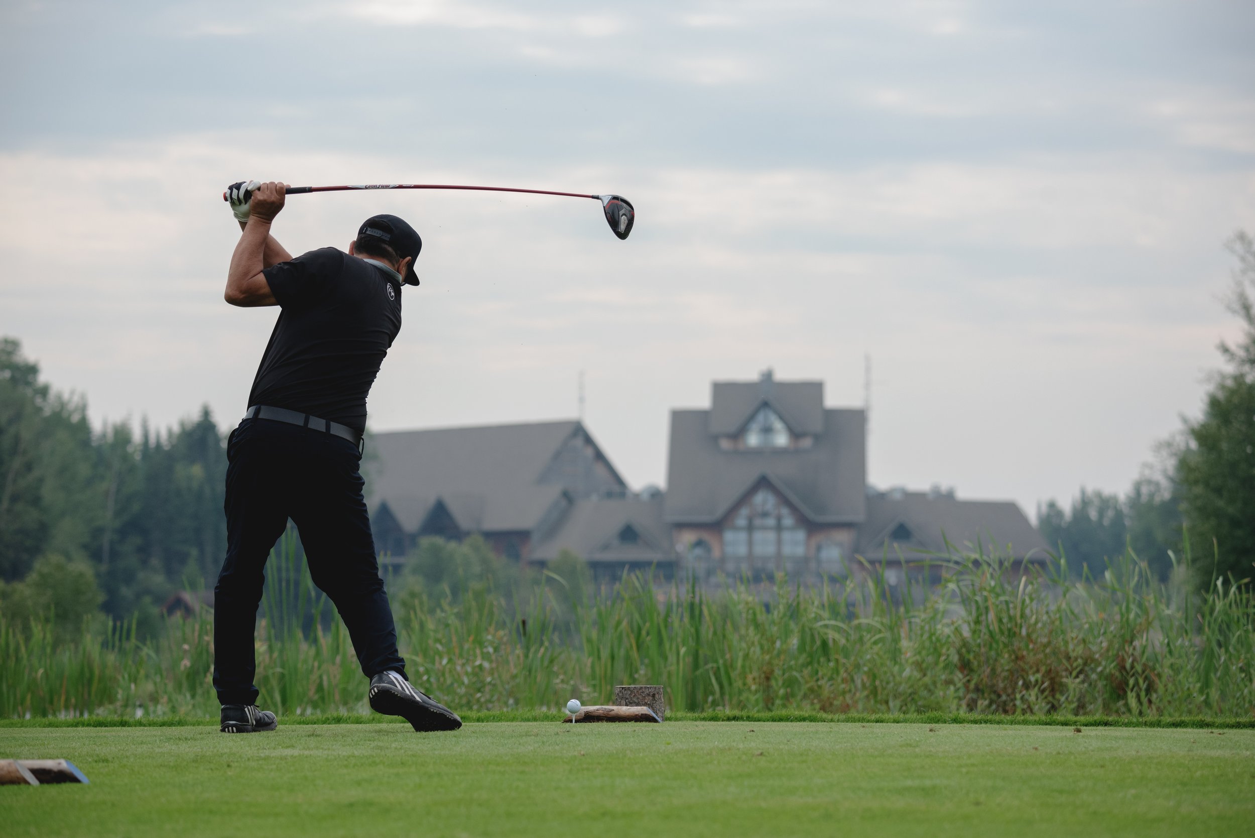 A man in golf attire swings a golf club on a golf course with a house and trees in the background.