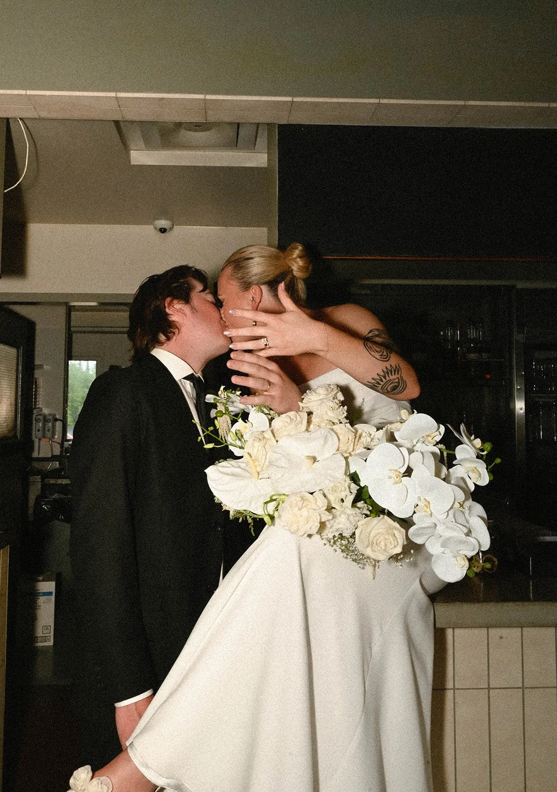 A bride and groom share a kiss at their wedding reception, with the bride's arms around the groom and the groom holding a large bouquet of white flowers.