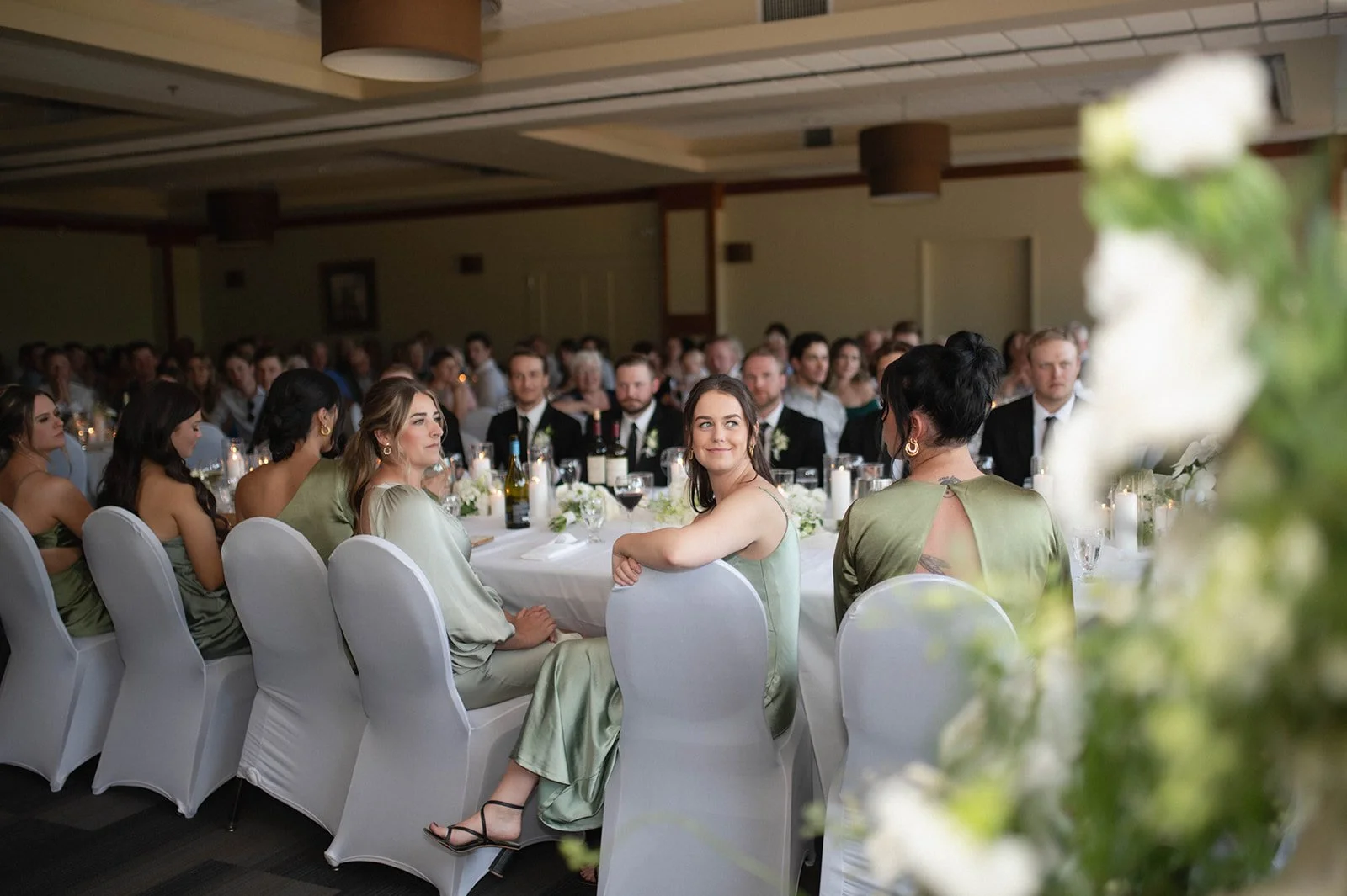 A group of people sitting at a formal event or banquet in a large room. Some are wearing formal attire, and there are candles and floral centerpieces on the table.