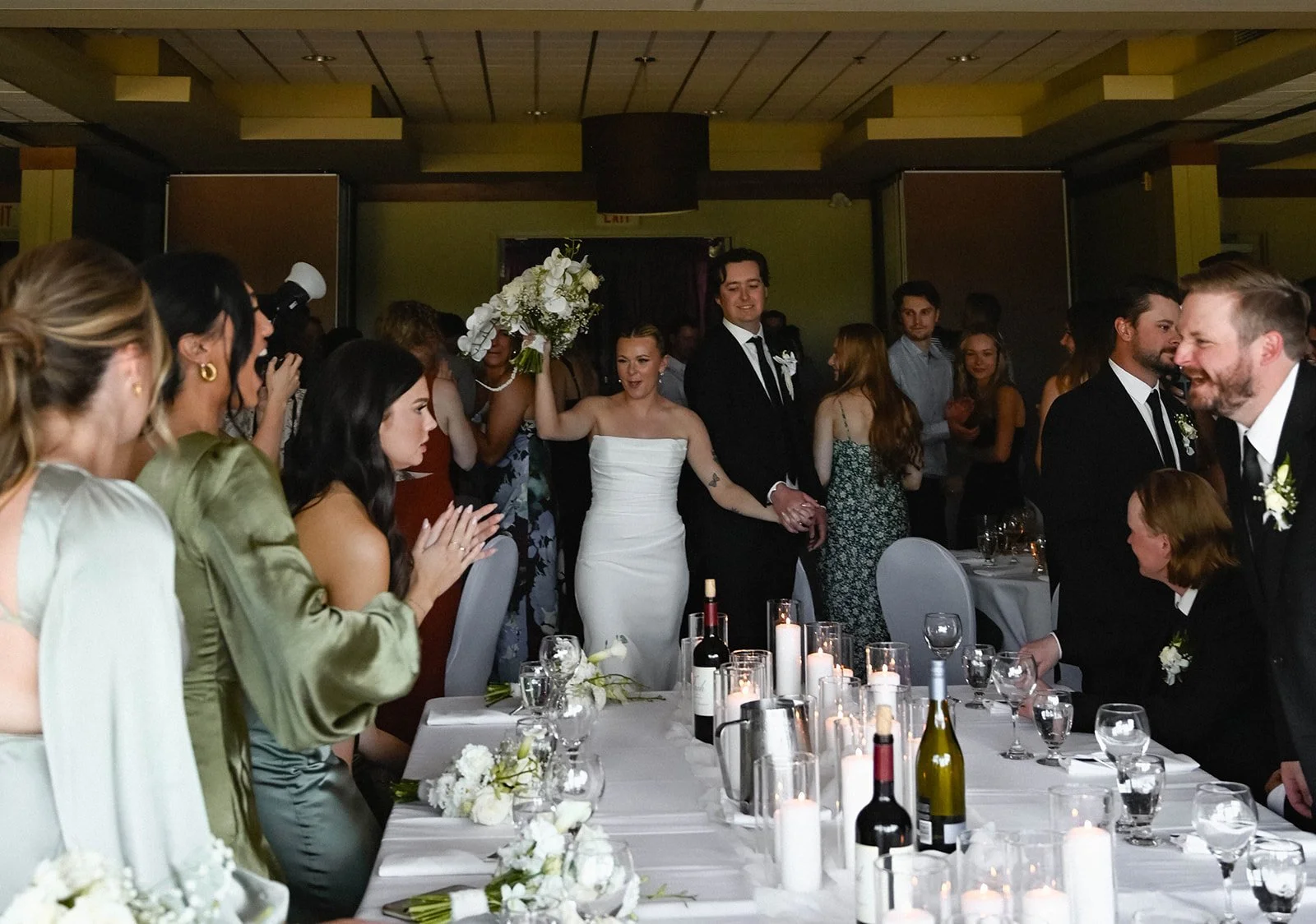 A wedding reception with the bride and groom holding hands and celebrating, surrounded by guests. The table is decorated with candles, wine bottles, and flowers.