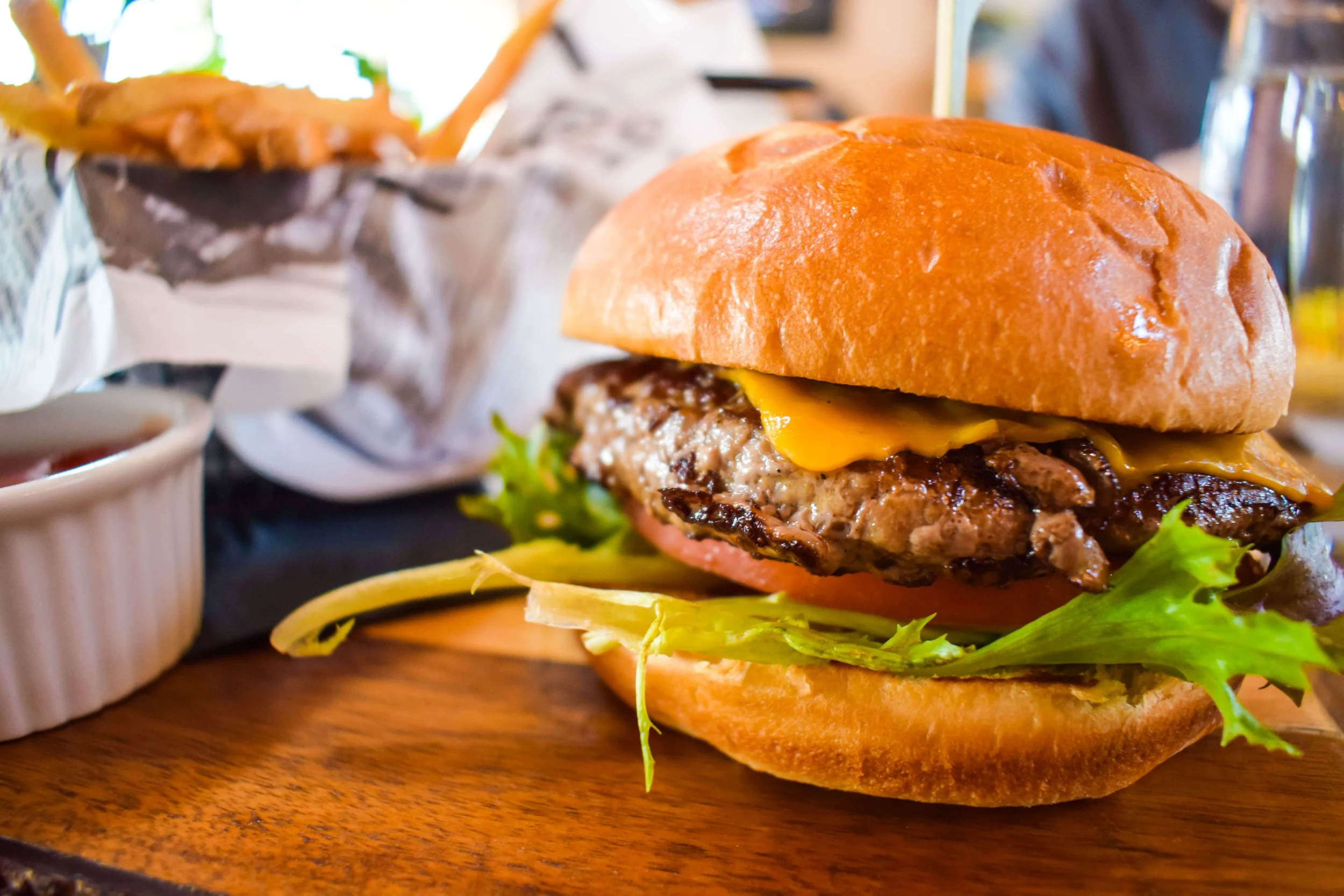 Close-up of a cheeseburger with a beef patty, melted cheese, lettuce, tomato, and a toasted bun. In the background, fries and condiment cups are visible.