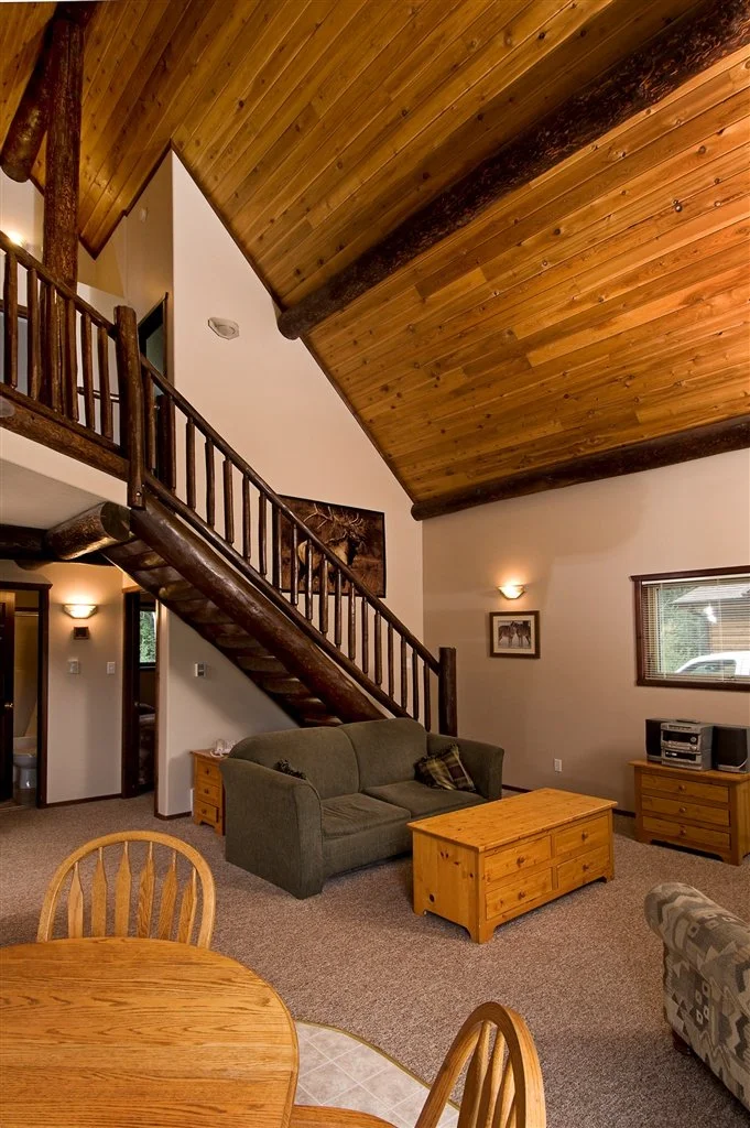 Living room with wood-paneled vaulted ceiling, wooden staircase with log railing, green sofa, wooden coffee table, and a small stereo on a side table.