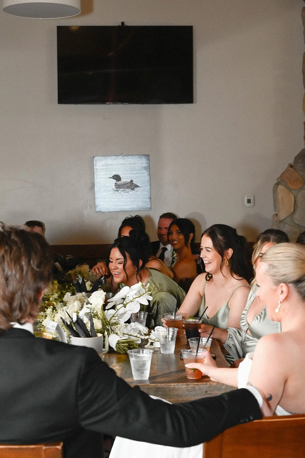People sitting at a decorated table, smiling and enjoying drinks, with a white wall and artwork of a duck in the background.