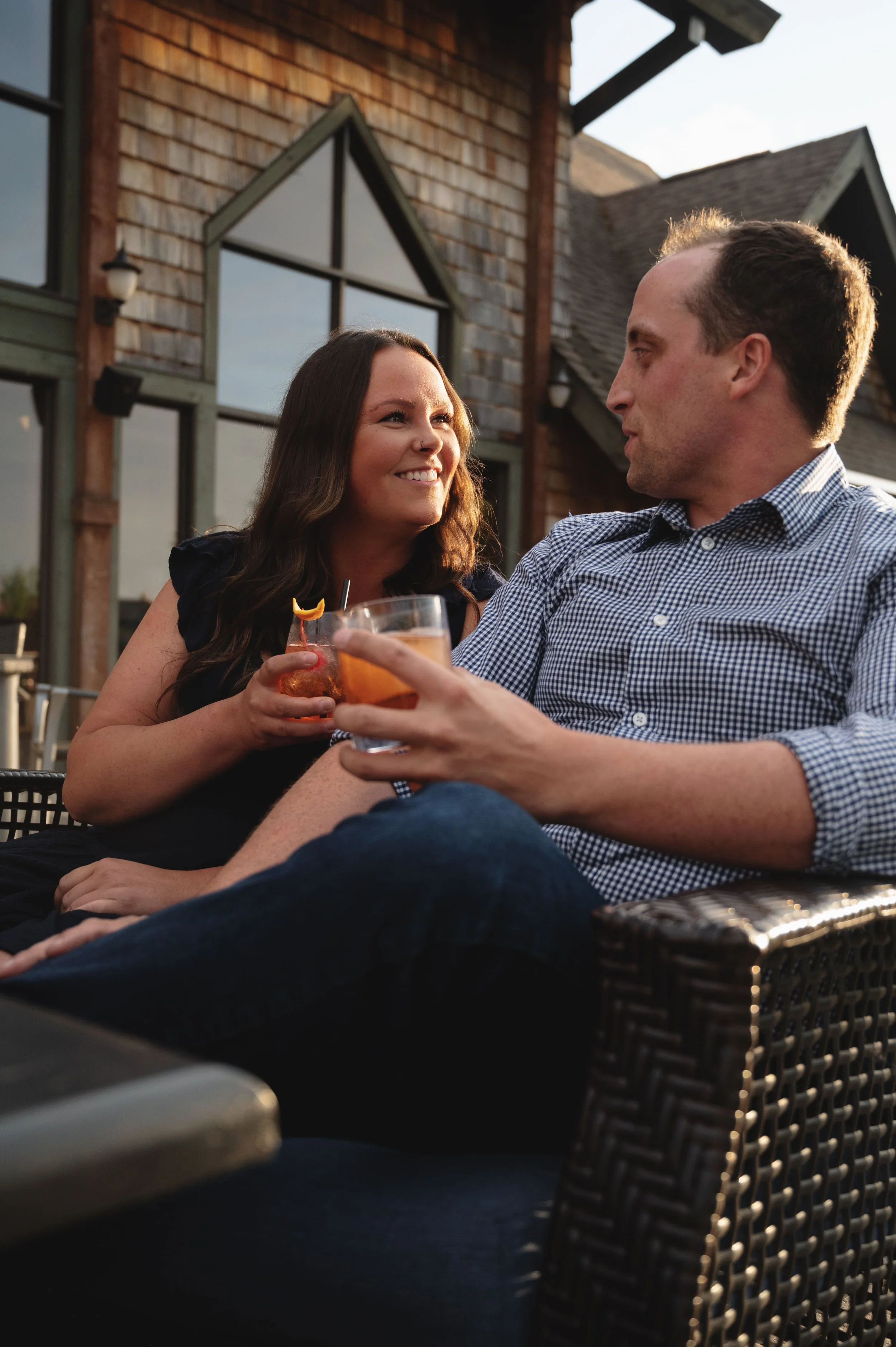 A man and a woman sitting outdoors, holding drinks and smiling at each other. The woman has long dark hair and is smiling at the man, who has short hair and is wearing a checkered shirt. In the background, there is a house with wooden siding and large windows, suggesting a social gathering on a balcony or patio during the daytime.