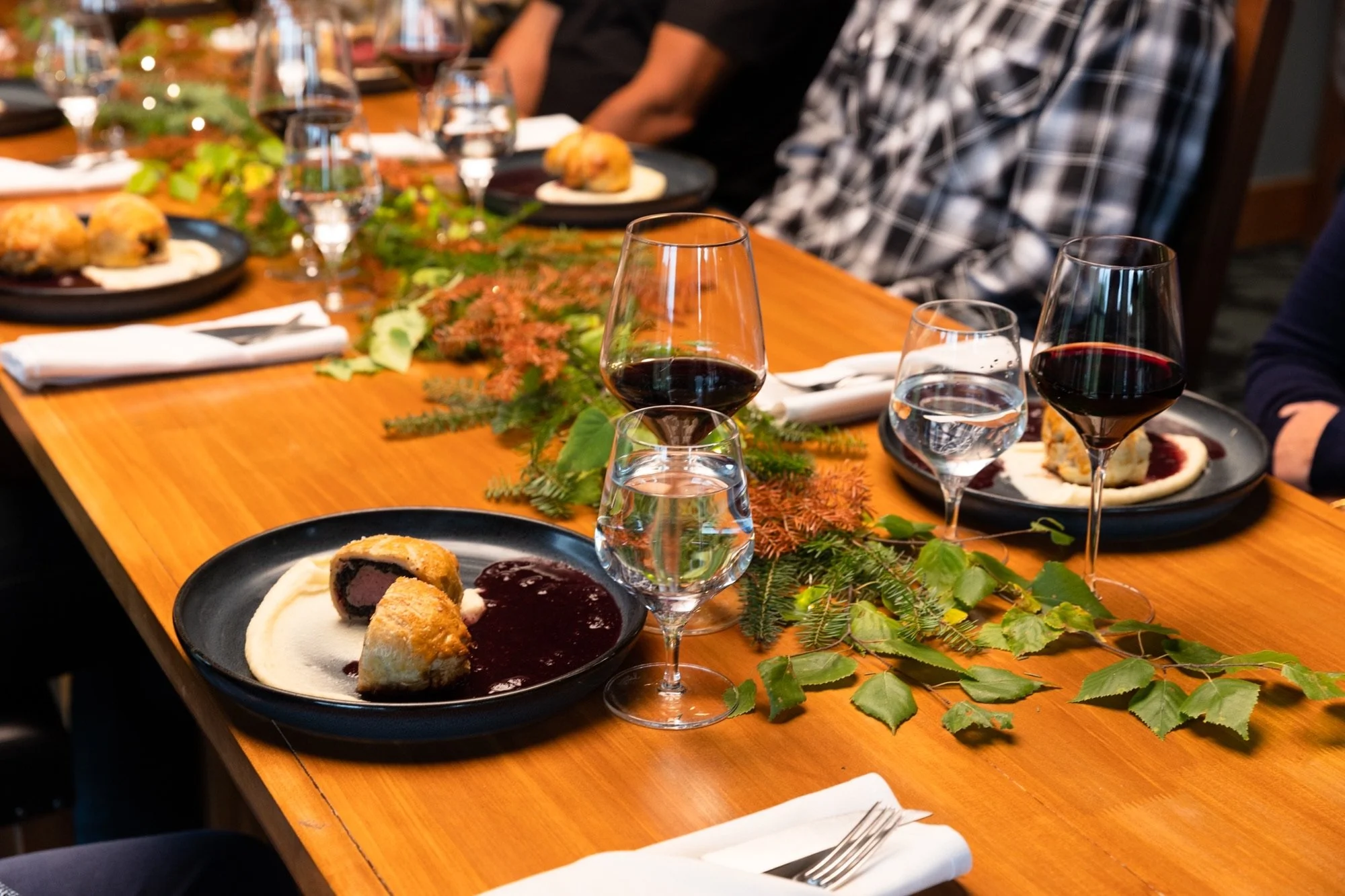 A holiday dinner table decorated with green foliage, glasses of red wine and water, and black plates with food including stuffed pork tenderloin with mashed potatoes and berry sauce, and pastry desserts.