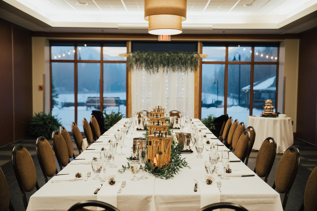 Decorated indoor wedding or event room with a long table set with glassware, lantern centerpieces, and pinecone accents, large windows with winter landscape outside, and a cake table in the corner.