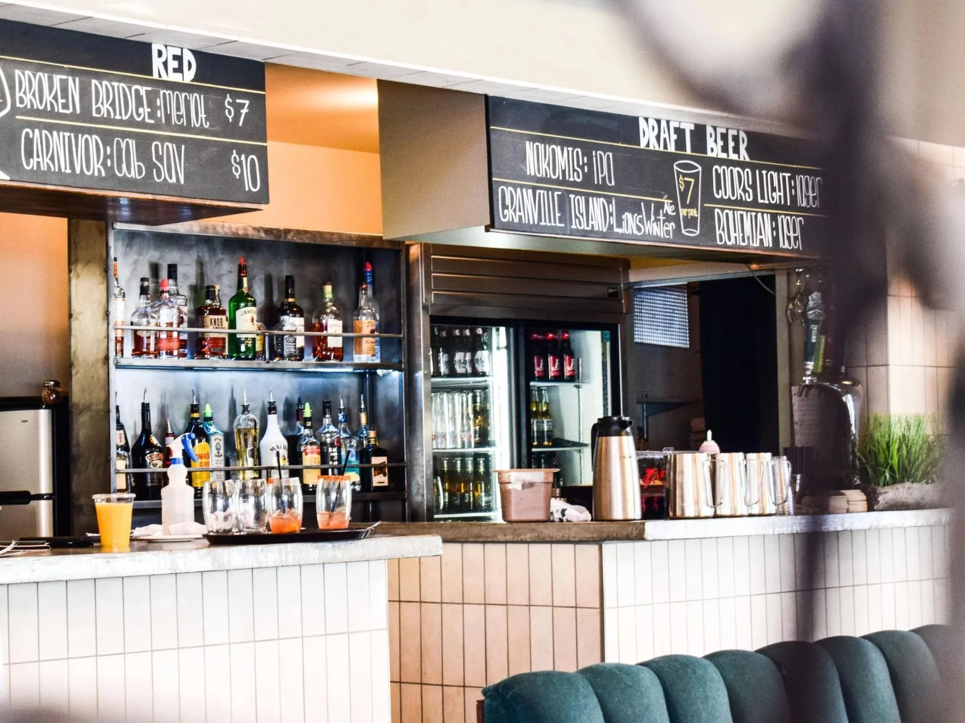 Bar area with bottles of liquor on shelves, a blackboard menu listing draft beer prices, a counter with drinks and cups, and some chairs in the foreground.