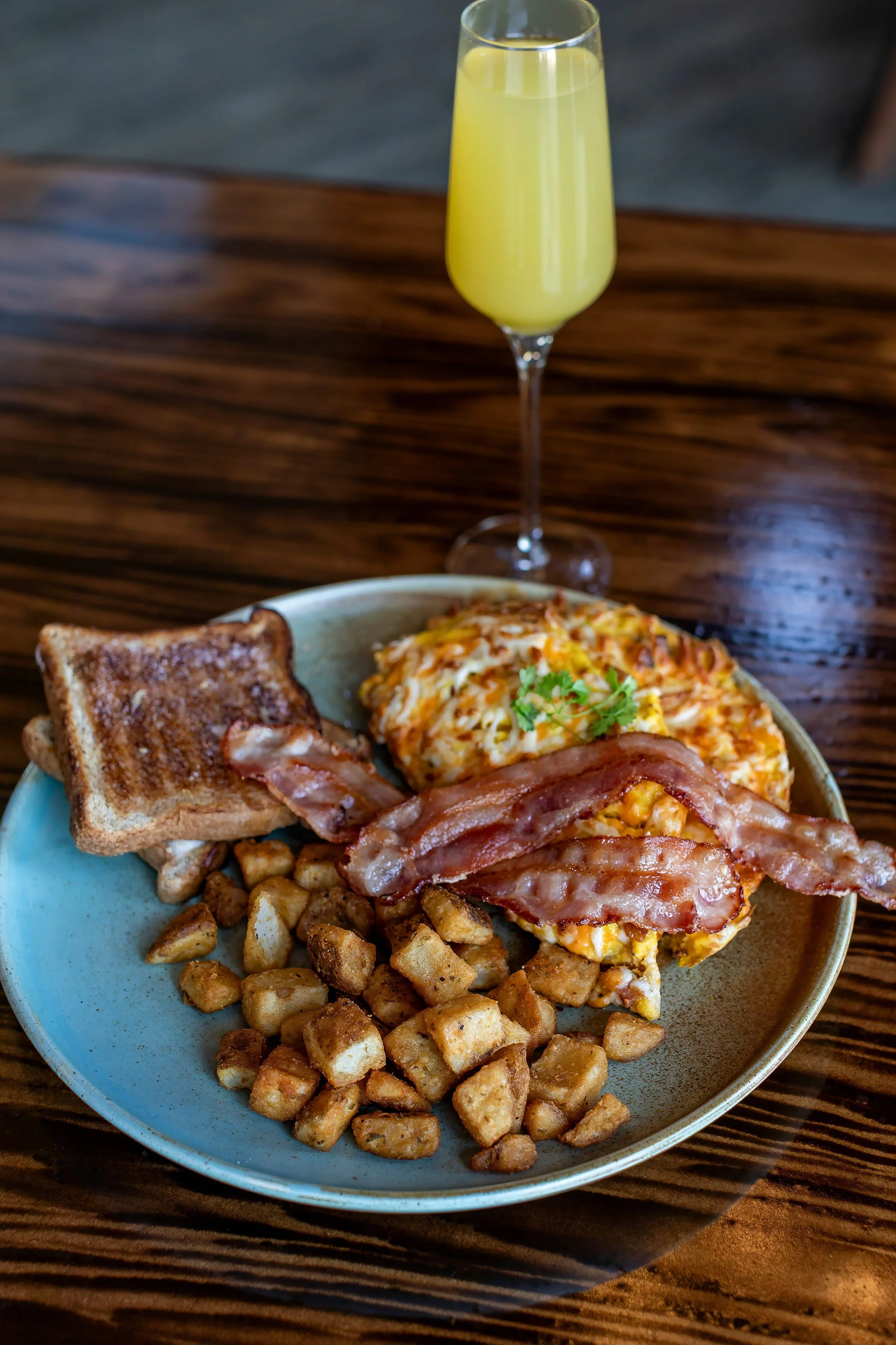 A breakfast plate with diced fried potatoes, two slices of crispy cooked bacon, scrambled eggs with cheese and herbs, and toasted bread, with a glass of yellow drink in the background on a wooden table.