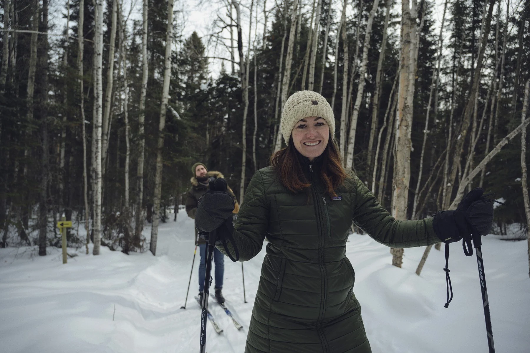 Two women cross-country skiing in a snowy forest, with one smiling and posing for the photo in the foreground and the other in the background.