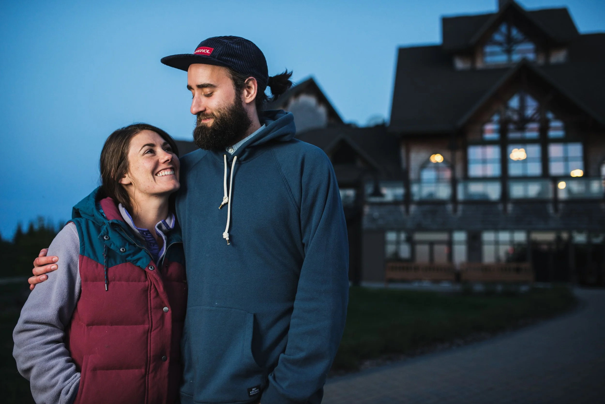 A smiling woman and a bearded man are outdoors at dusk, embracing each other in front of a large house with illuminated windows.