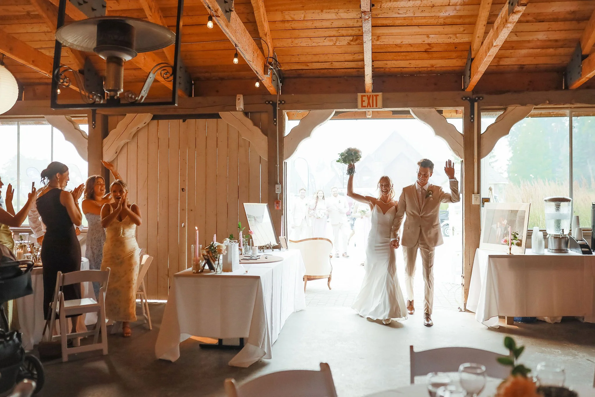 A wedding reception scene with a bride and groom smiling and waving as they enter a rustic venue with wooden beams. Guests are clapping and celebrating inside, and the entrance has large open doors with bright natural light outside.