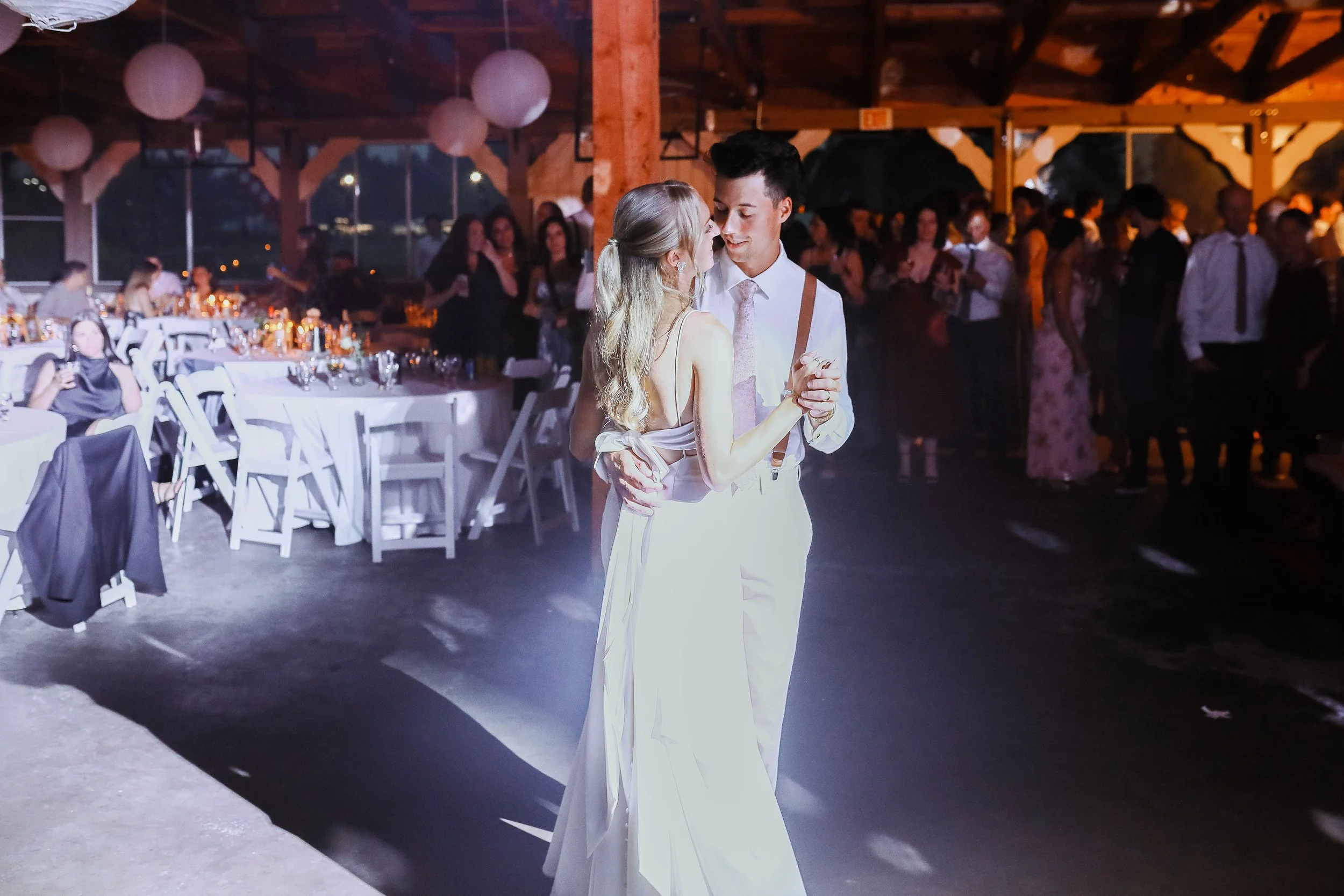 A couple dancing at a wedding reception, with guests in the background, decorated tables, and hanging paper lanterns in a barn-like venue.