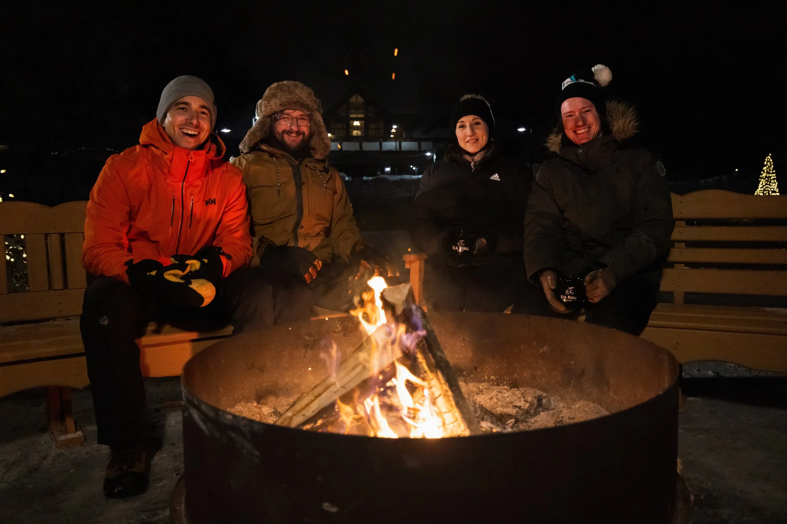 Four people sitting around a campfire at night, wearing winter jackets and hats, with a house and a lit Christmas tree in the background.