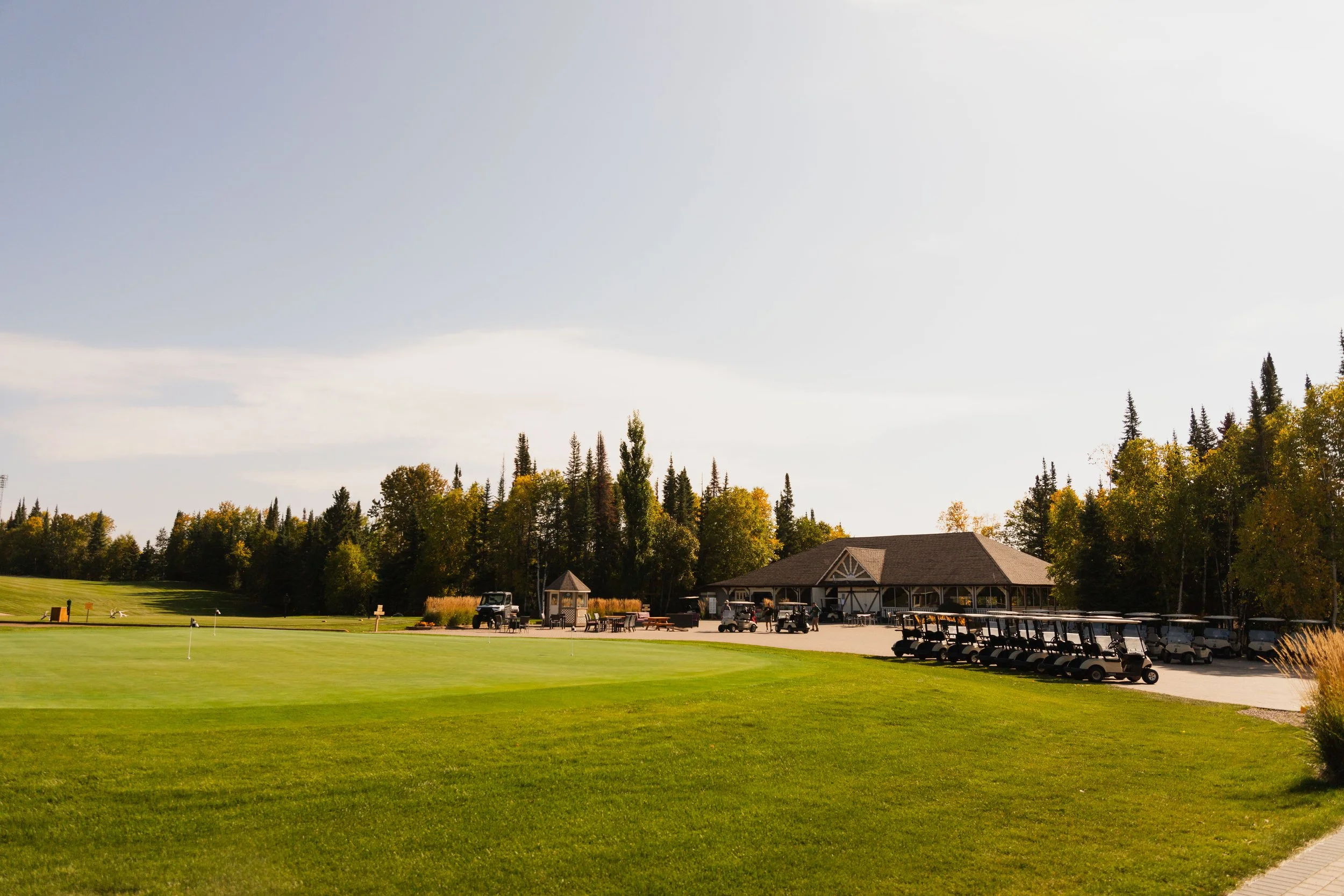 A golf course with a putting green in the foreground, a clubhouse and golf carts in the background, surrounded by tall trees under a partly cloudy sky.