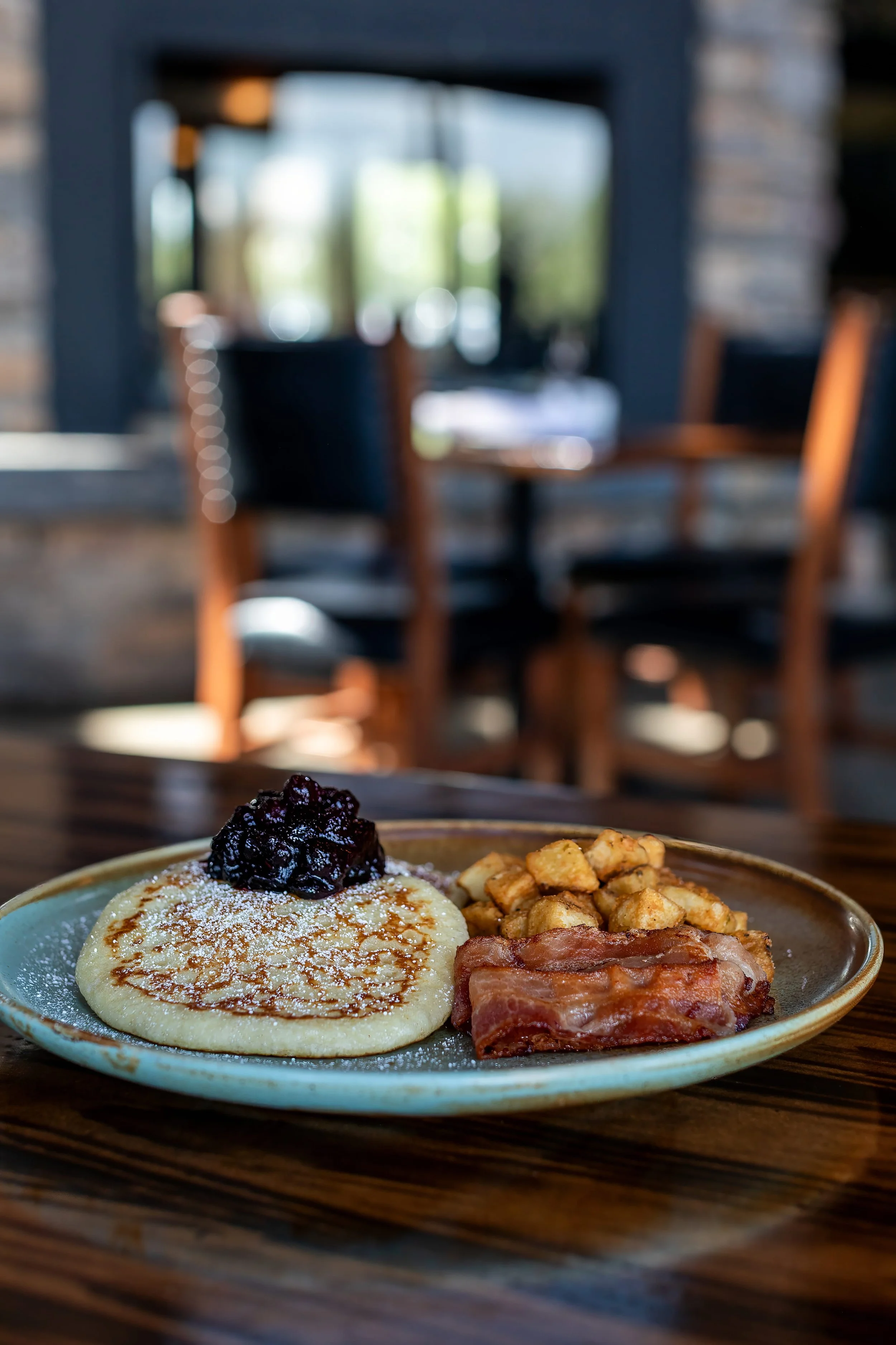 A plate of breakfast food including a pancake with a dollop of blueberry jam and powdered sugar, breakfast meat, diced potatoes, and bacon, on a wooden table in a cozy restaurant setting.
