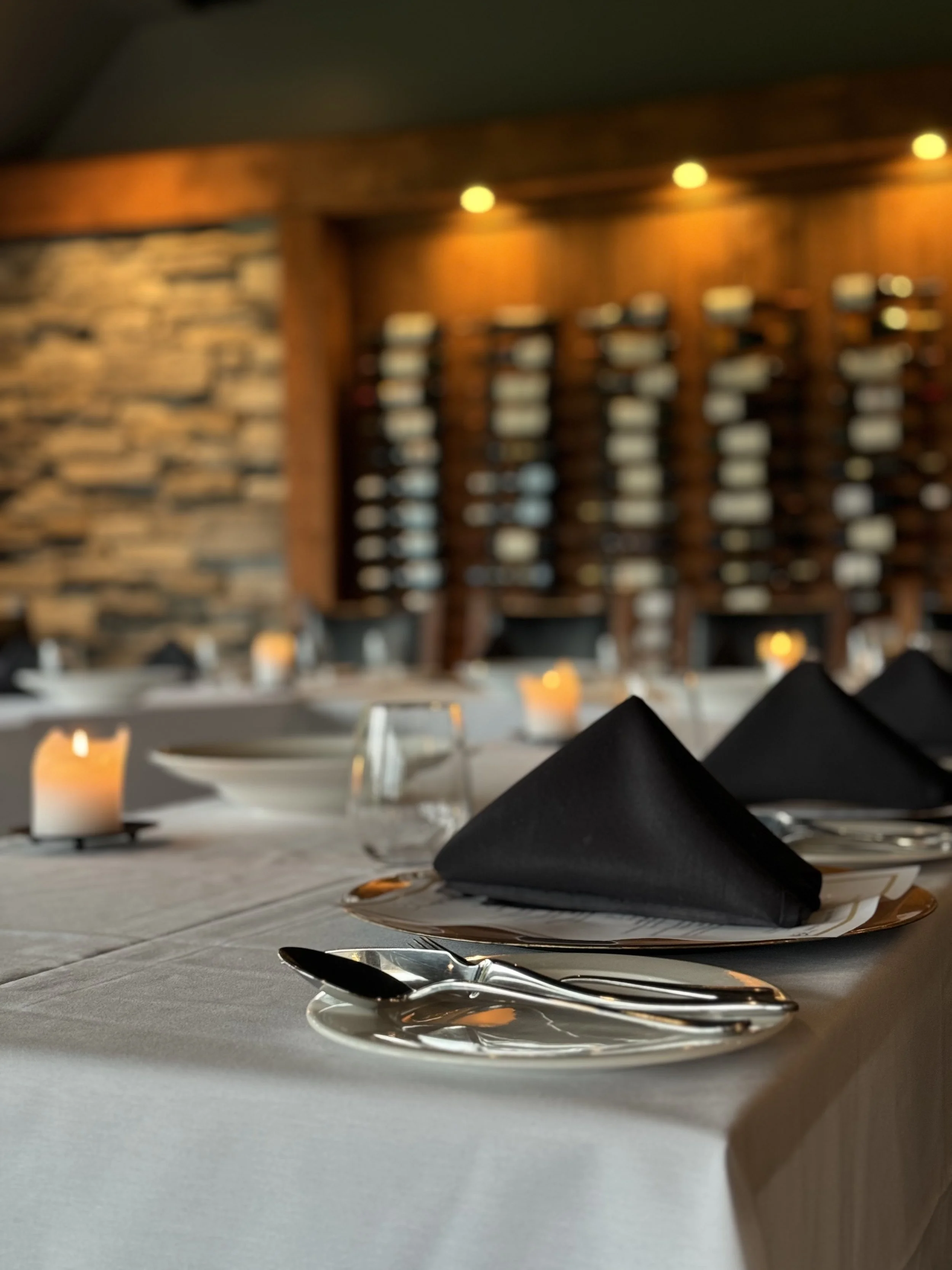 A decorated dining table with black napkins folded into triangles, placed on white plates, and silverware, against a background of a brick and wood-paneled wall with warm lighting.