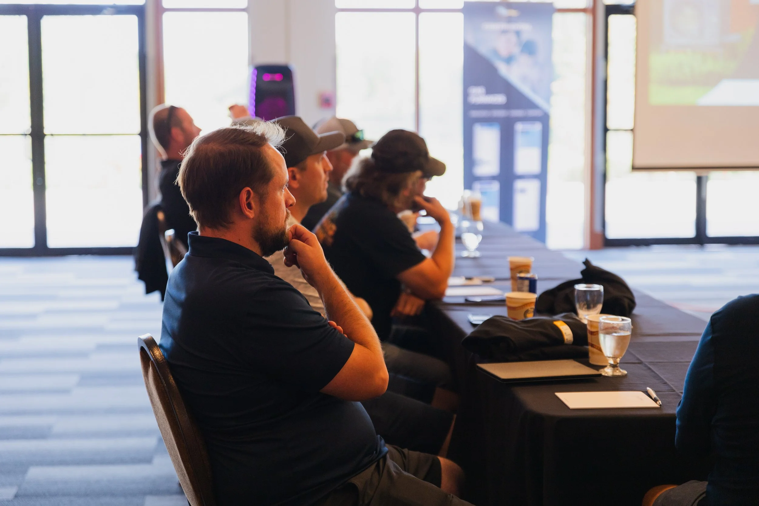 A group of people sitting at a long table during a meeting or conference, with drinks and notepads in front of them, in a room with large windows letting in natural light.