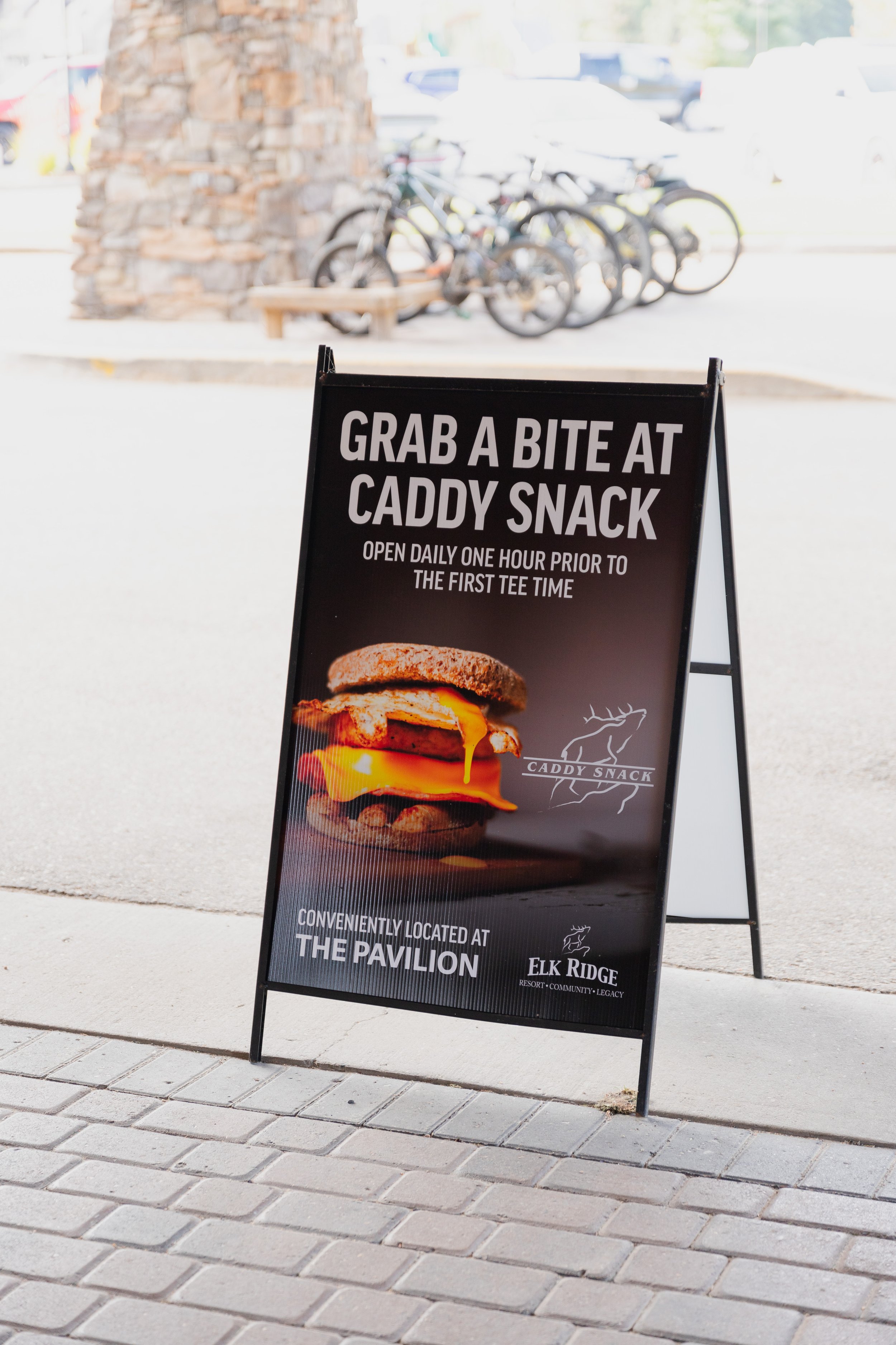 Outdoor sidewalk sign advertising a food establishment called Caddy Snack, with a hamburger image and text about location at Elk Ridge Resort.